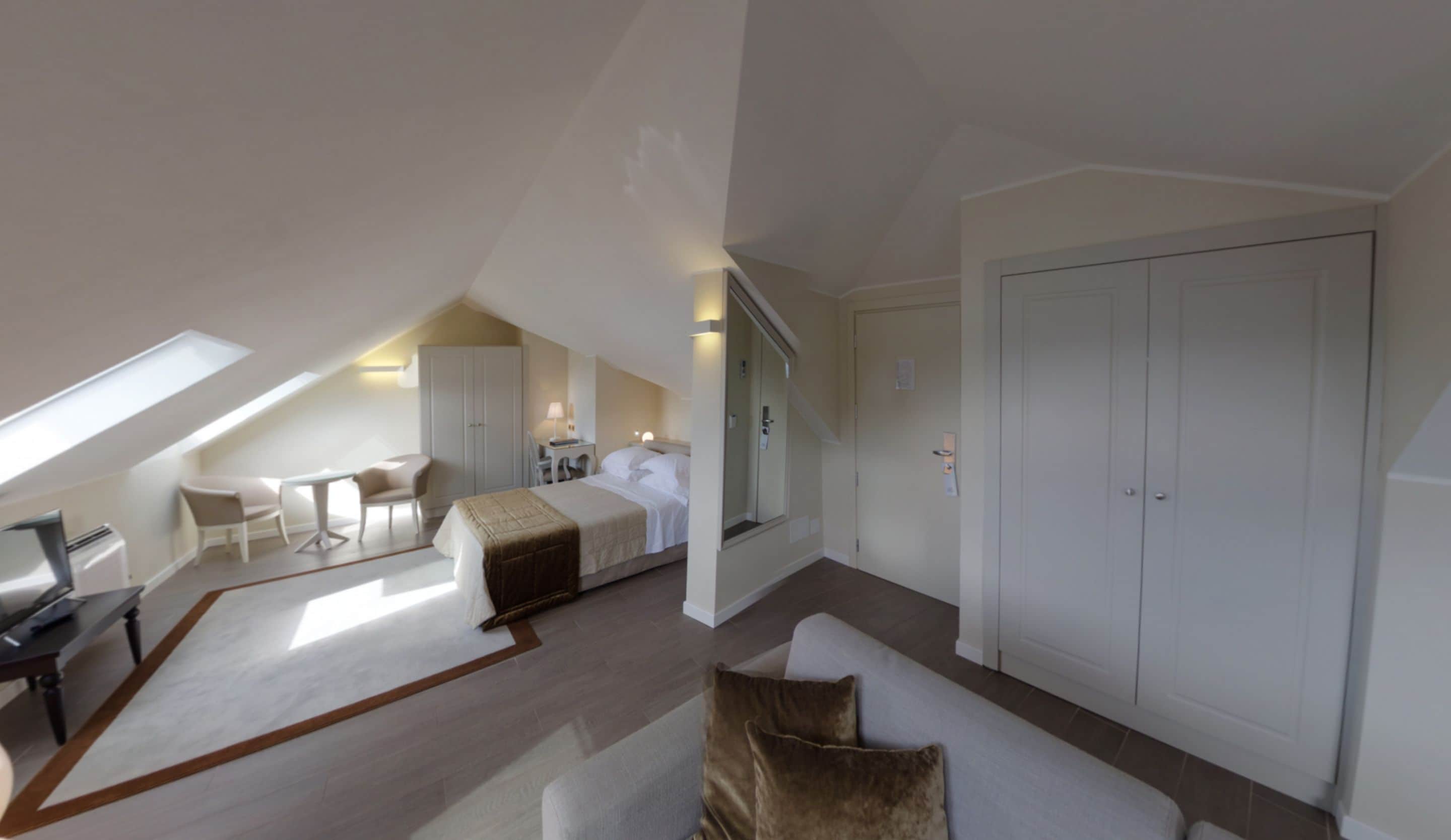 Una habitación de hotel moderna con techo inclinado, una cama grande, una zona de estar con mesa y sillas, un armario y un espejo amplio.