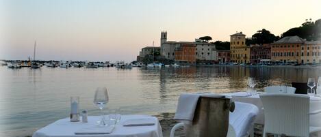 A waterfront dining area with tables set for two, overlooking a marina with boats and buildings.