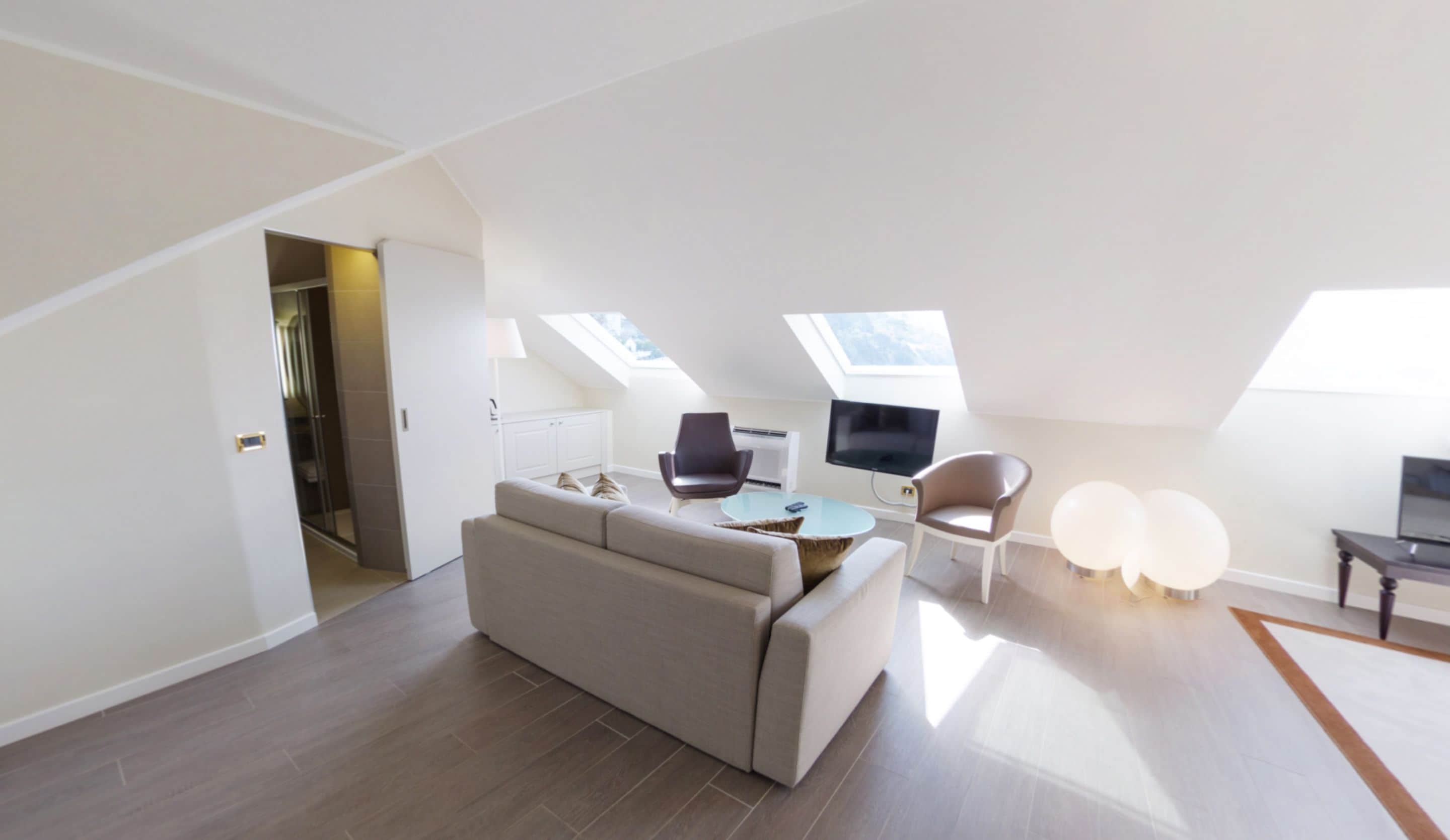 A modern loft apartment with a high ceiling, skylights, a sofa, two armchairs, a coffee table, a flat-screen TV, and a small table with a lamp.