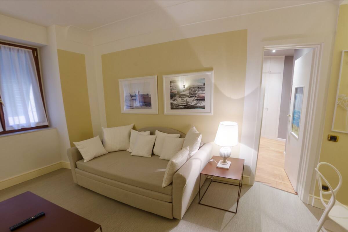 Een moderne hotelkamer met een beige bank, twee ingelijste foto’s aan de muur, een klein nachtkastje met een lamp, en een deur naar een andere kamer.