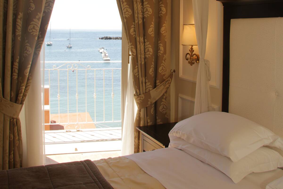 Een hotelkamer met een bed, gordijnen, uitzicht op zee en een balkon.