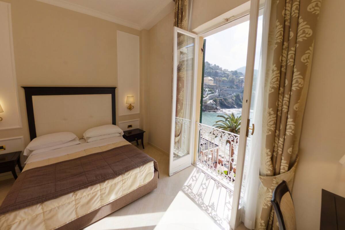 Een slaapkamer met een bed, nachtlampjes, een nachtkastje en een balkon met uitzicht op zee.