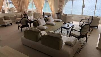 A spacious living room with a large sofa, armchairs, and a coffee table, featuring a view of the sea and a mountainous landscape.