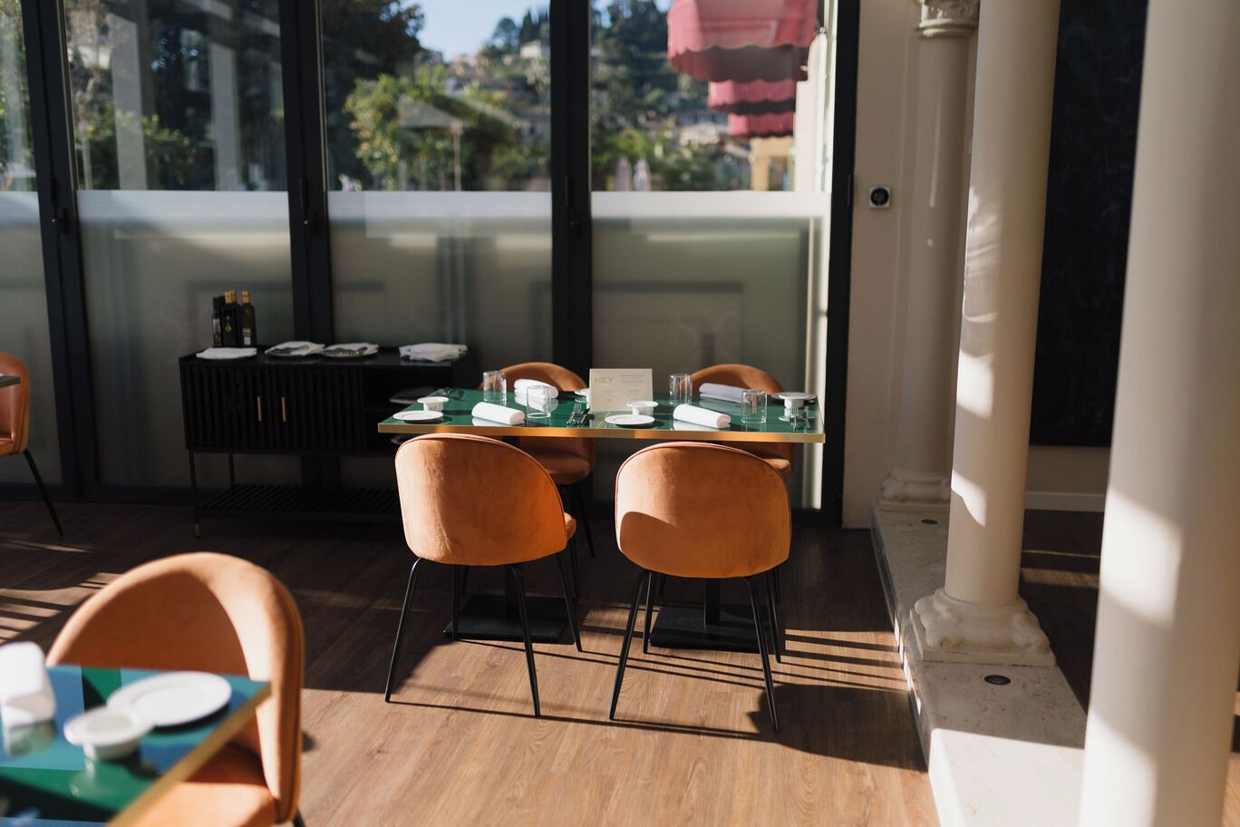 A dining area with a glass table, orange chairs, and a view of buildings through large windows.