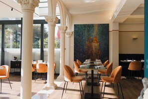 A modern dining area with a large table and orange chairs, surrounded by white columns and large windows.