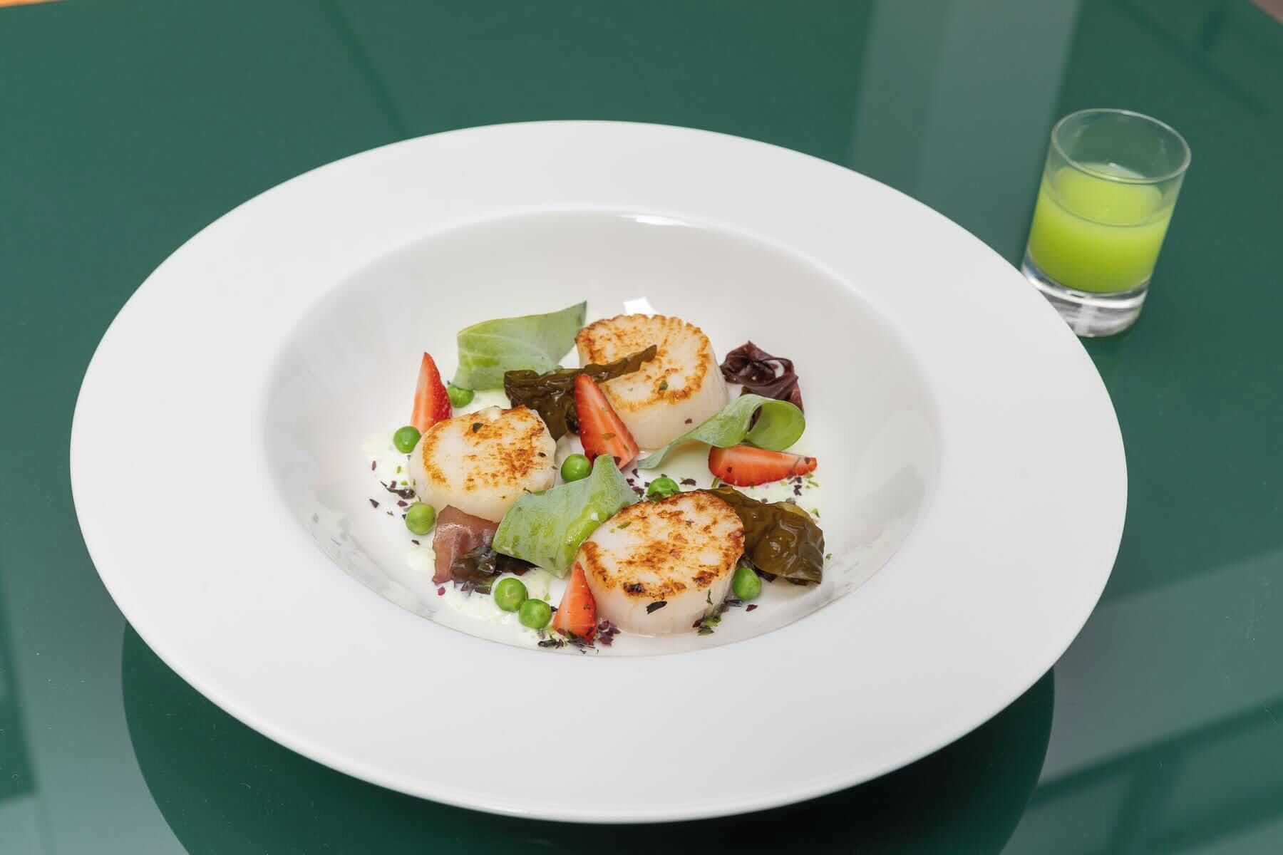 Un plato blanco con vieiras selladas, verduras y una bebida verde sobre una mesa verde.