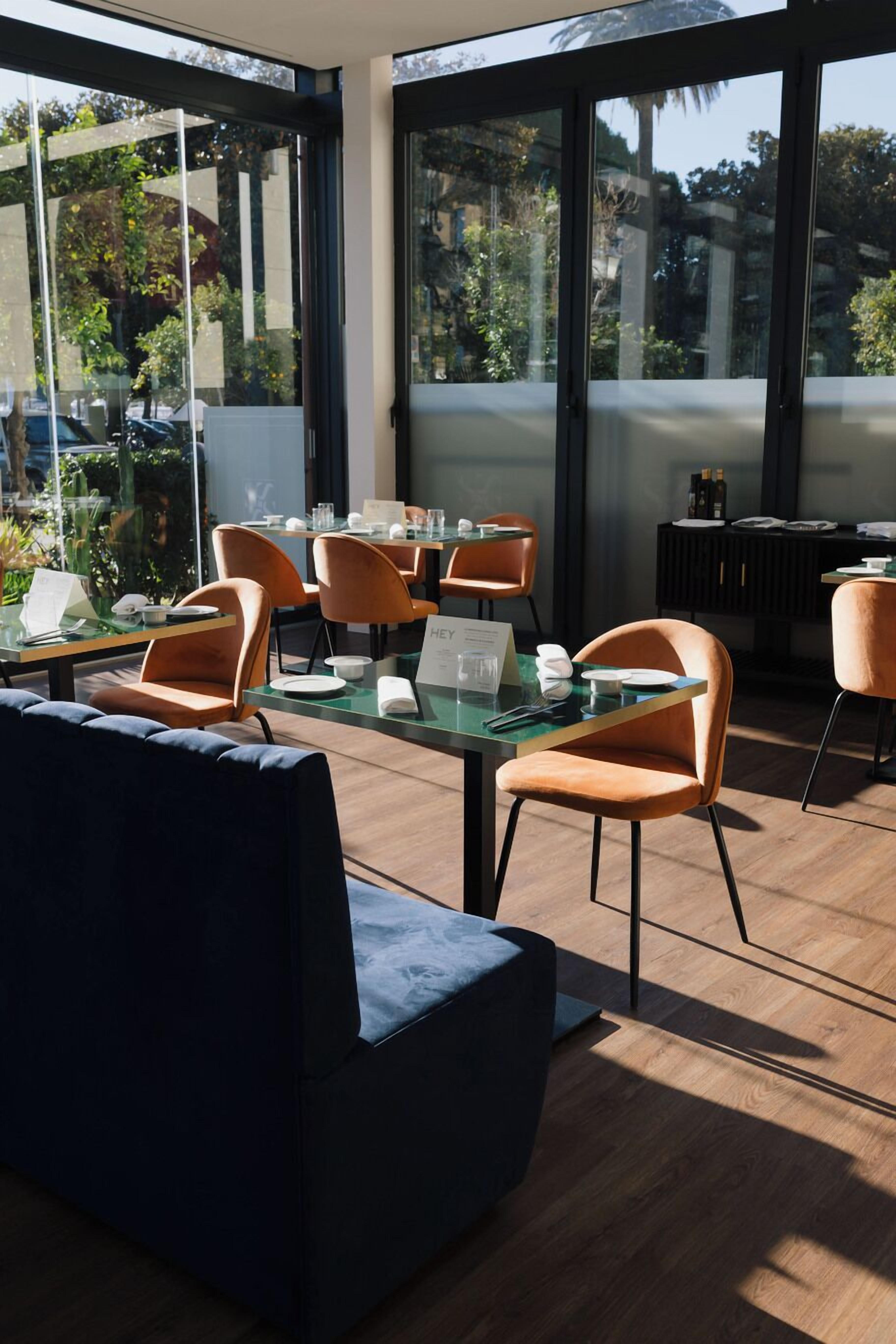 Un espace repas moderne avec des tables en verre et des chaises orange, un canapé bleu foncé et une vue sur la verdure à l’extérieur.