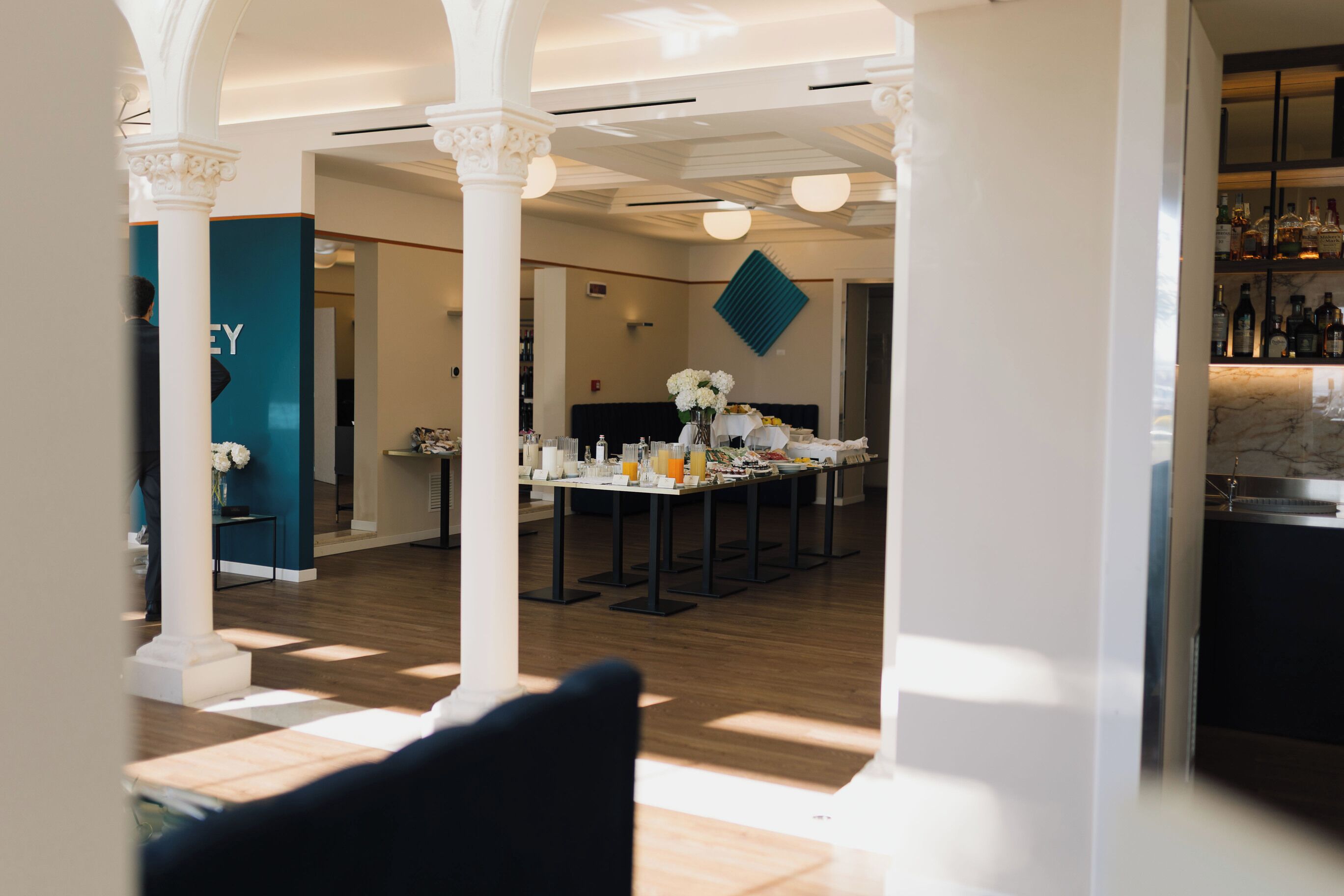 En modern hotellobby med ett centralt placerat matområde, vita pelare och en bar med flaskor.