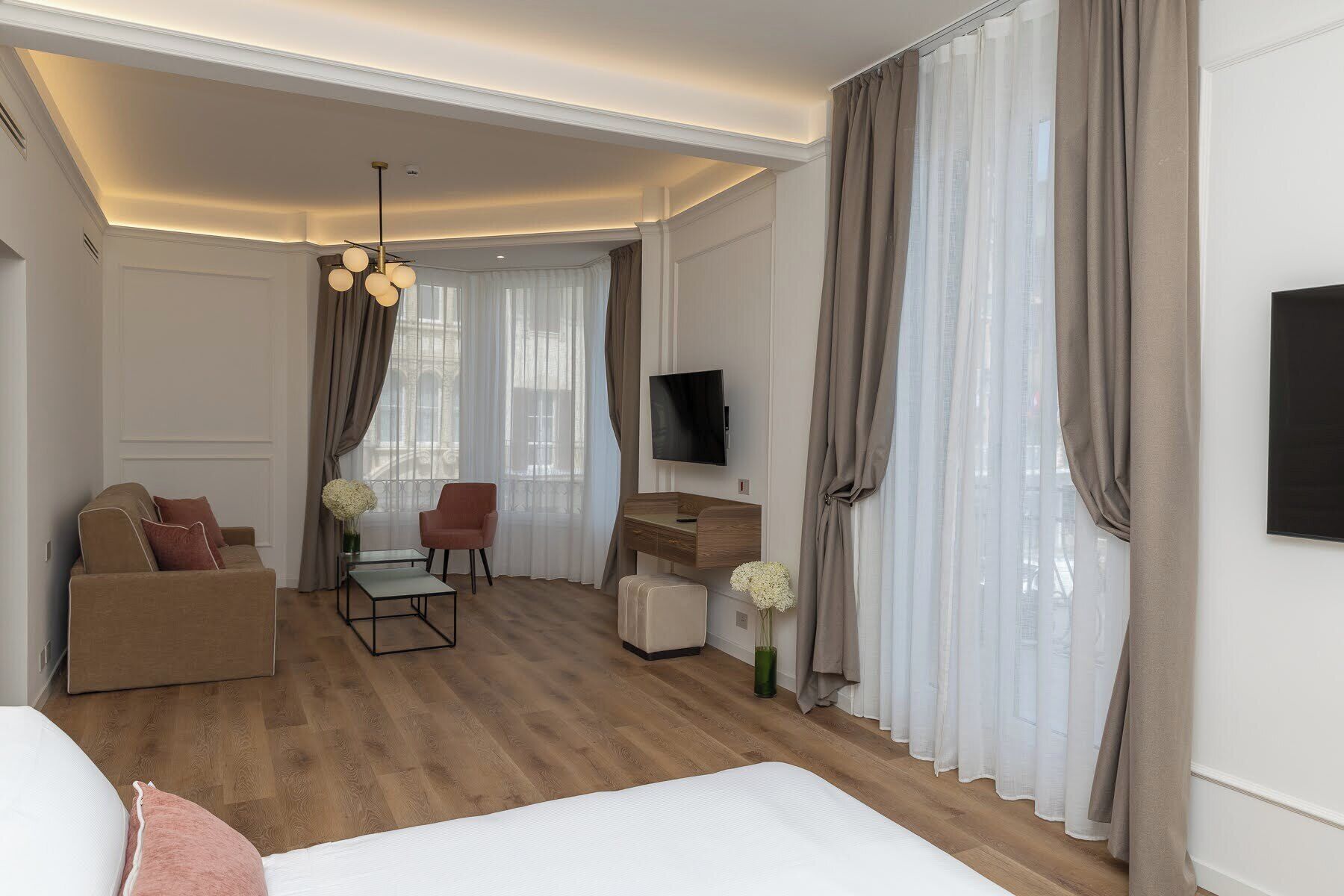 Habitación de hotel con cama, sofá, escritorio, silla, televisor y un ventanal con cortinas.