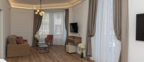 Habitación de hotel con cama, sofá, escritorio, silla, televisor y un ventanal con cortinas.