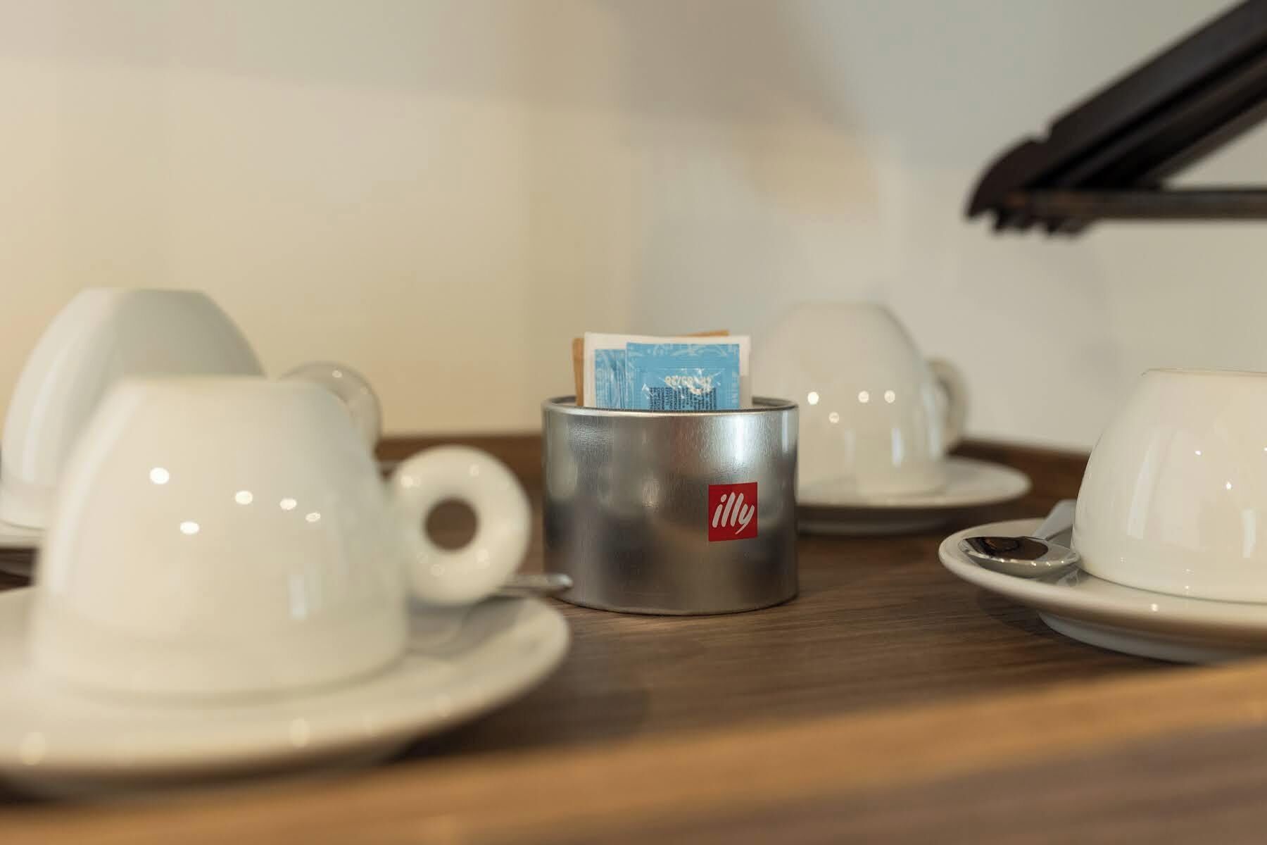 Un coin café avec une tasse en métal portant l’inscription « illy » et contenant une carte, entouré de tasses et de soucoupes blanches sur une table en bois.