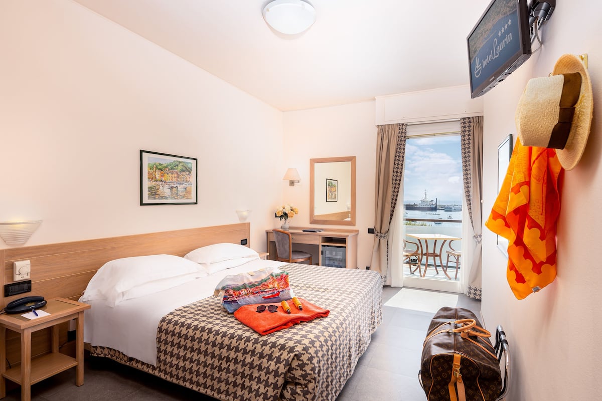 Une chambre d’hôtel avec un lit, un bureau, une chaise, une valise et une vue sur la mer.