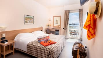 A hotel room with a bed, a desk, a chair, a suitcase, and a view of the sea.