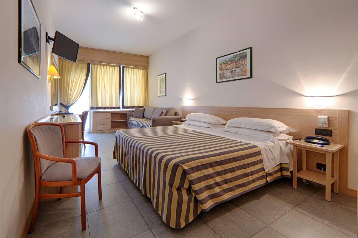 Une chambre d’hôtel comprenant un lit, un bureau, une chaise, une table de chevet, une télévision et une fenêtre avec des rideaux.