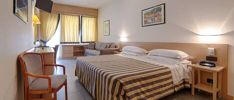 Habitación de hotel con cama, escritorio, silla, mesita de noche, televisor y ventana con cortinas.