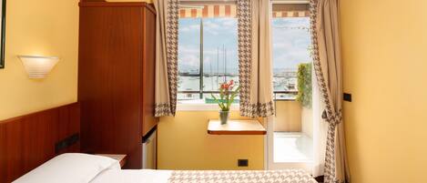 A hotel room with a bed, a nightstand, a window with curtains, a view of the marina, and a wall-mounted light fixture.