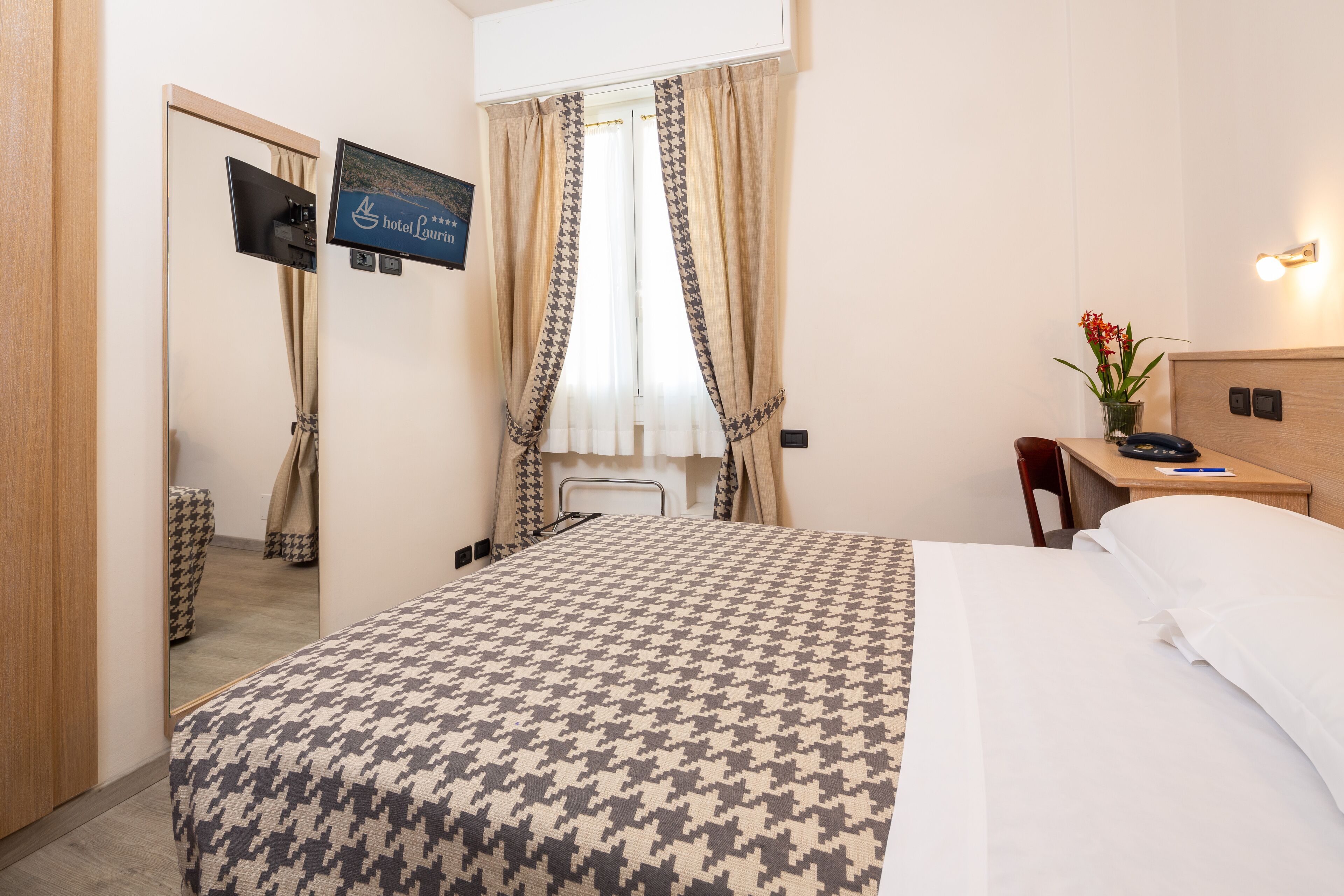 Una camera d'albergo con un letto, una scrivania, una sedia, una televisione e una finestra con tende.