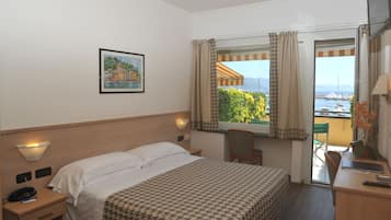 A hotel room with a bed, a desk, a chair, a window with curtains, a wall-mounted lamp, a painting, and a view of the sea and boats.