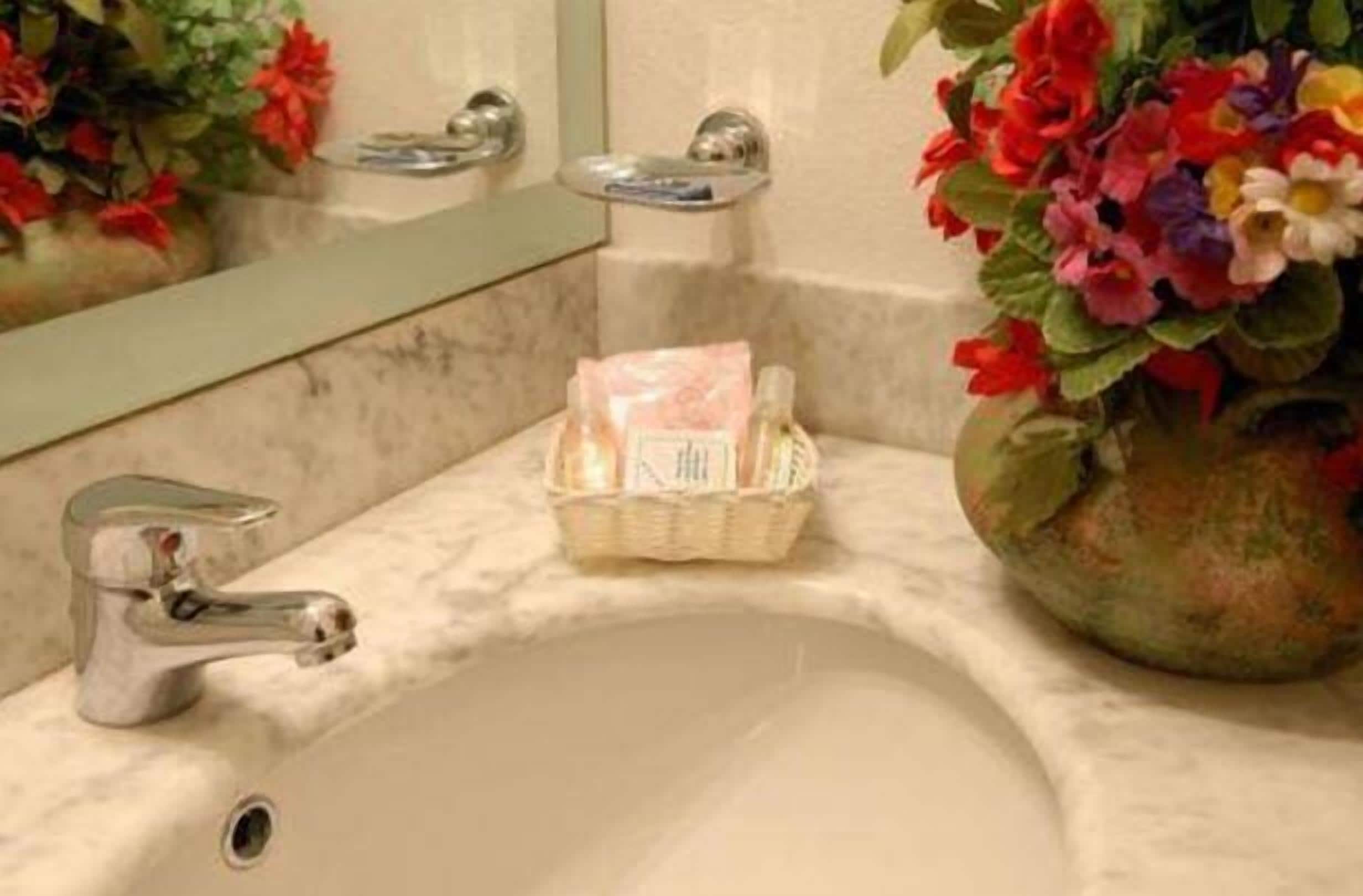 Uma pia de banheiro com bancada de mármore, um espelho, um vaso com flores e uma cesta com produtos de higiene pessoal.