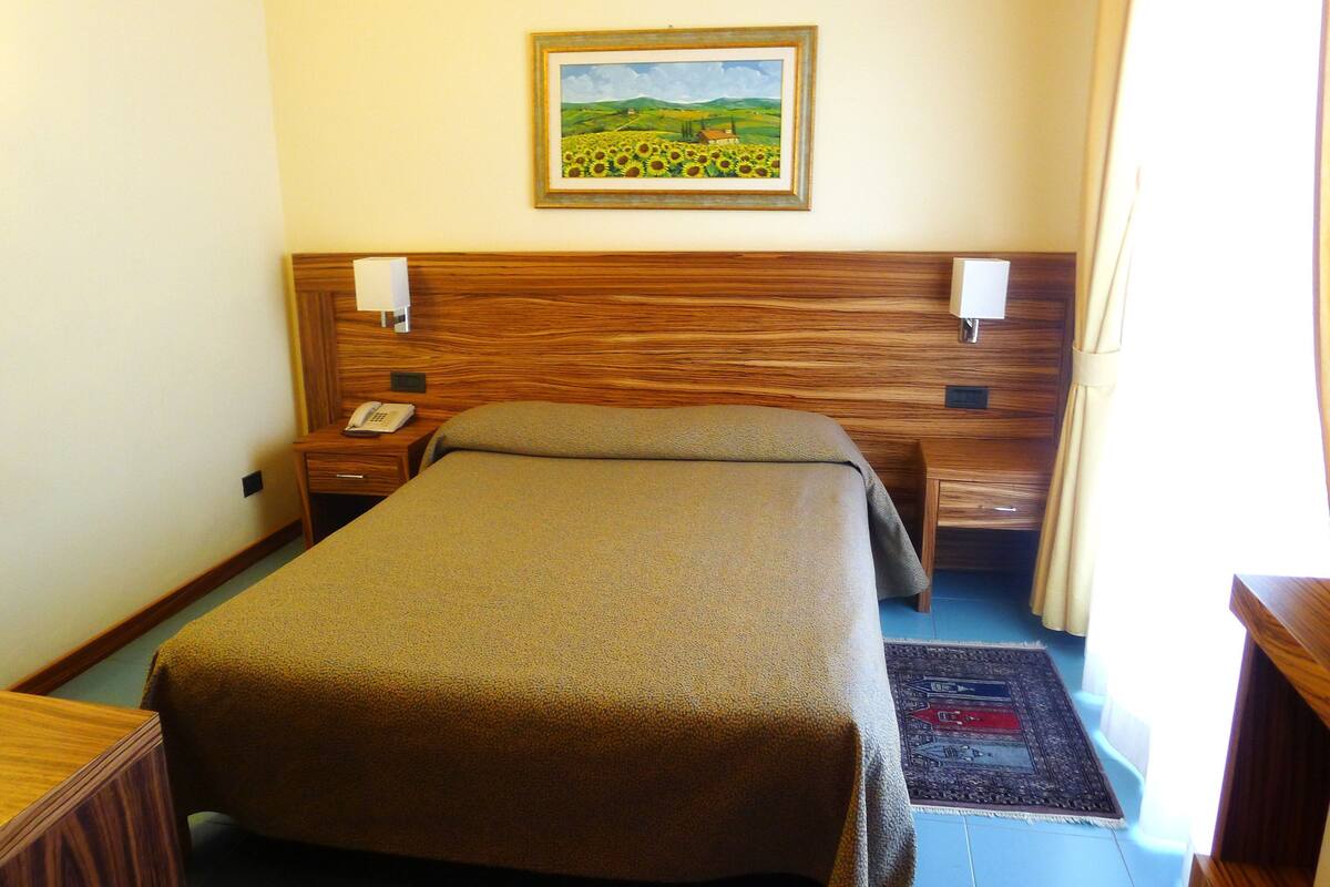A hotel room with a wooden headboard, a single bed, a bedside table with a phone, and a painting of a landscape.