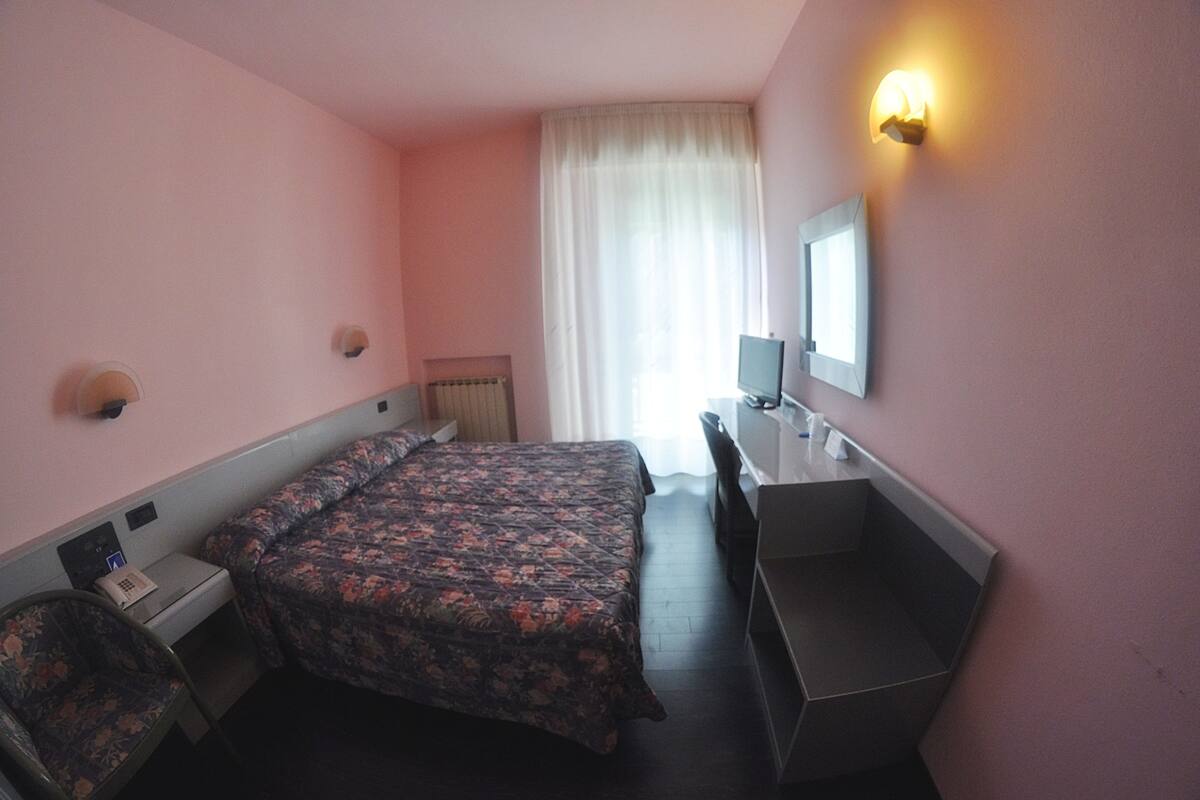 A hotel room with a bed, a desk, a chair, a mirror, and a window with curtains.