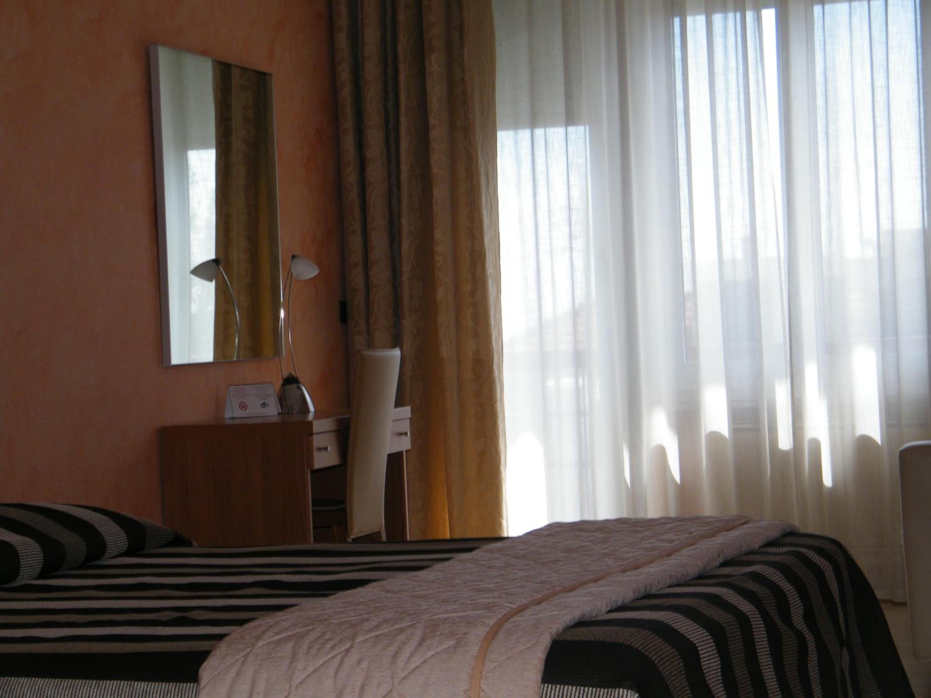 A hotel room with two beds, a desk with a lamp, a chair, and a window with sheer curtains.