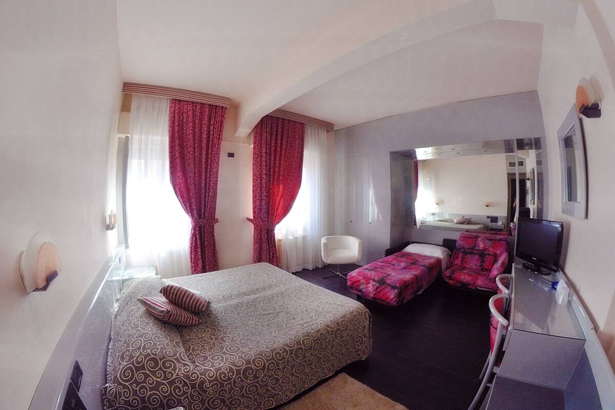 A hotel room with a bed, two bedside tables, a desk with a chair, a TV, and a bathroom visible through a door.