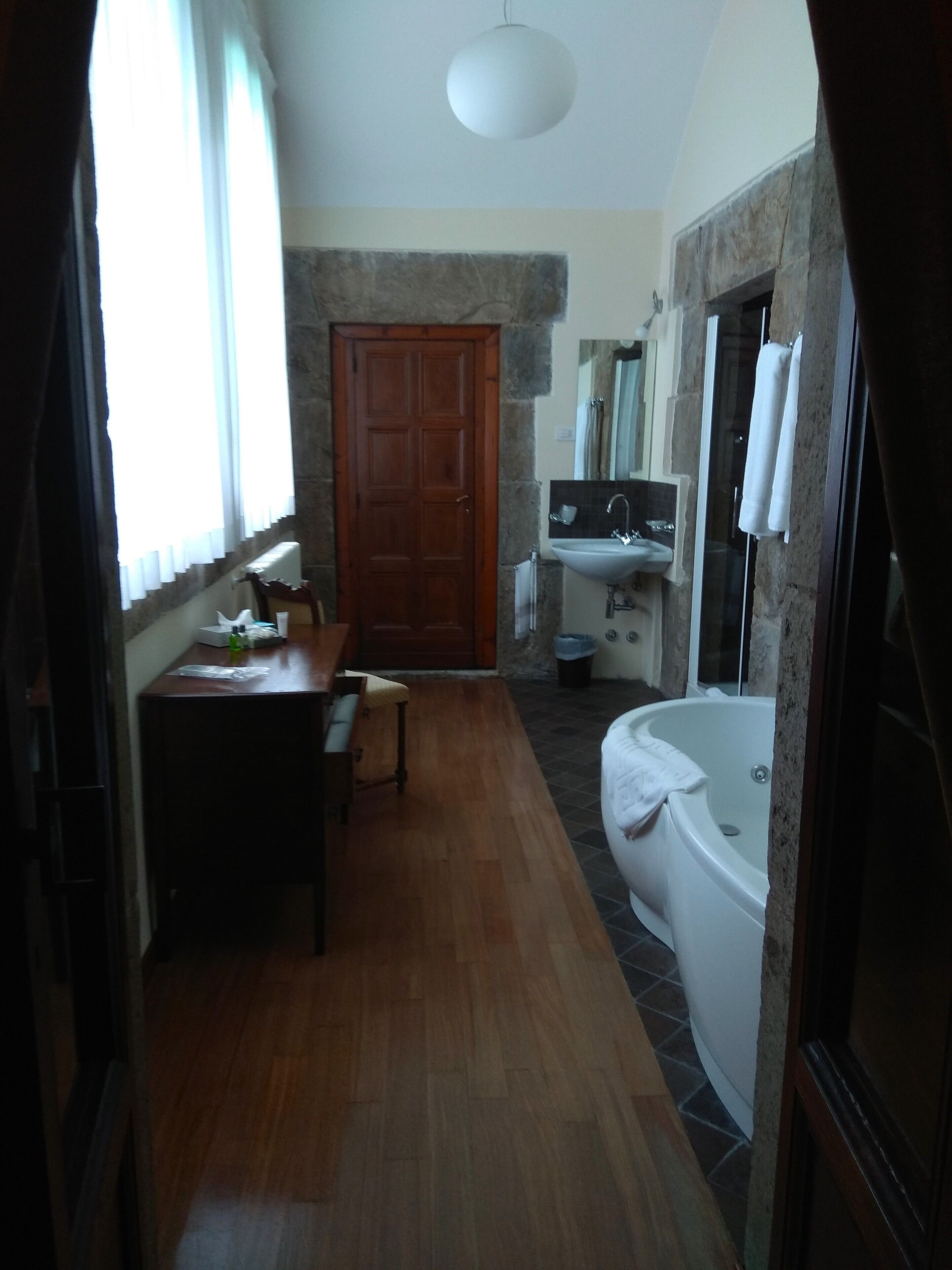 A narrow hallway with a wooden door, a small table with a chair, a white bathtub, and a bathroom with a sink and mirror.