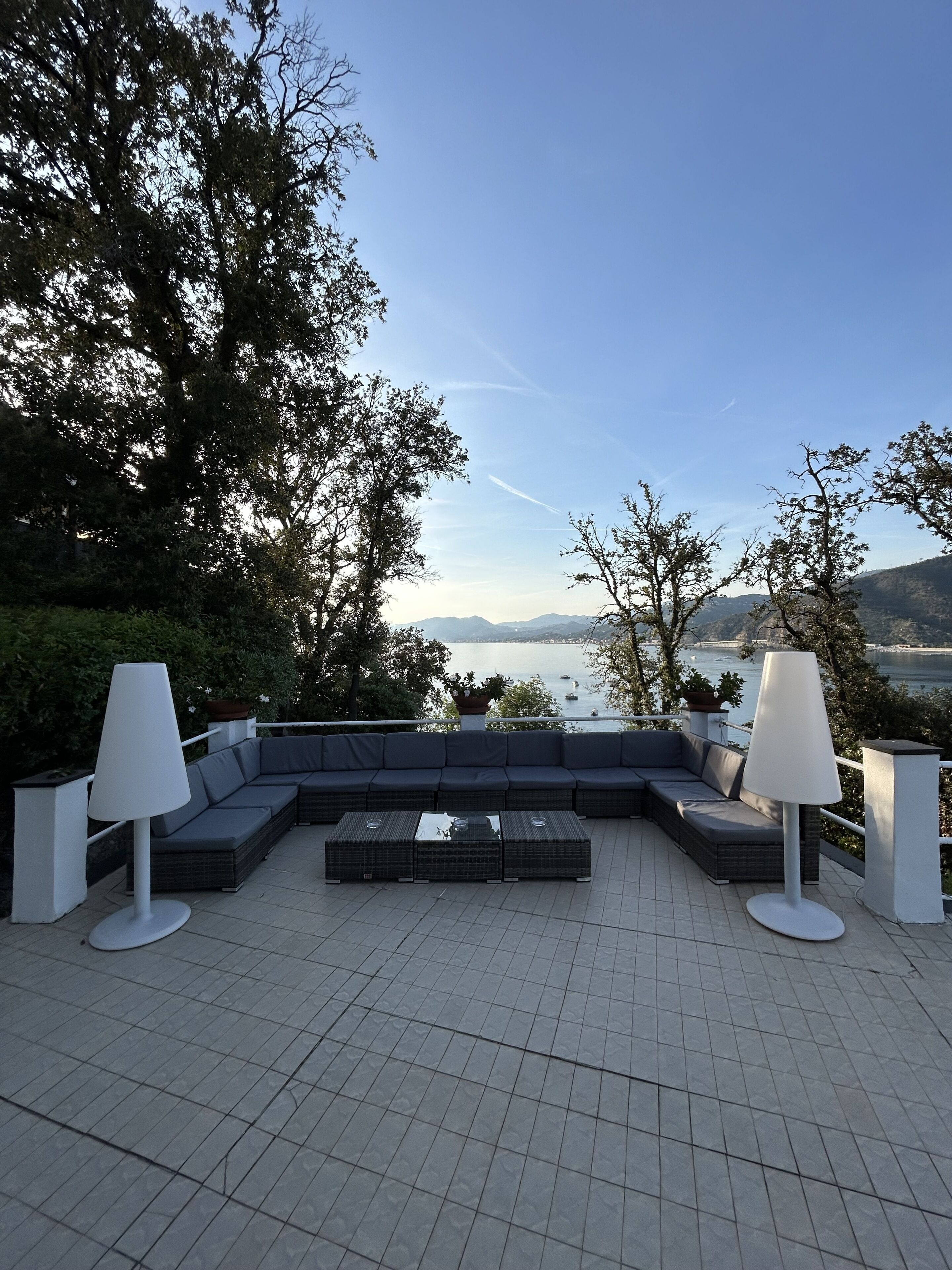 A terrace with a sectional sofa, two floor lamps, and a view of the water and mountains.