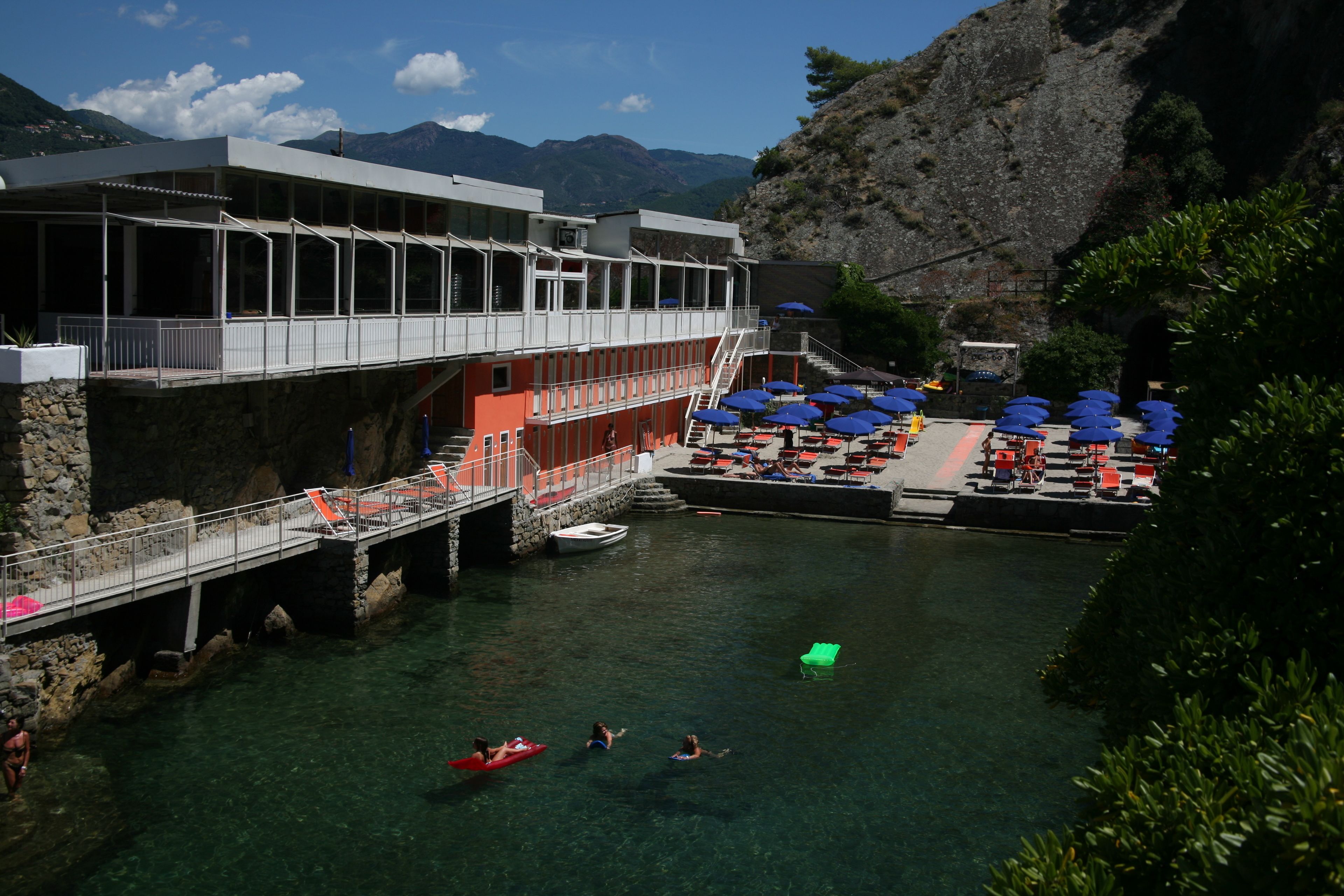 A waterfront hotel with a pool area, outdoor seating, and a view of the mountains.