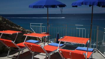 Sillas de playa con cojines azules y cubiertas rojas, dispuestas sobre una terraza de hormigón con vista al océano.