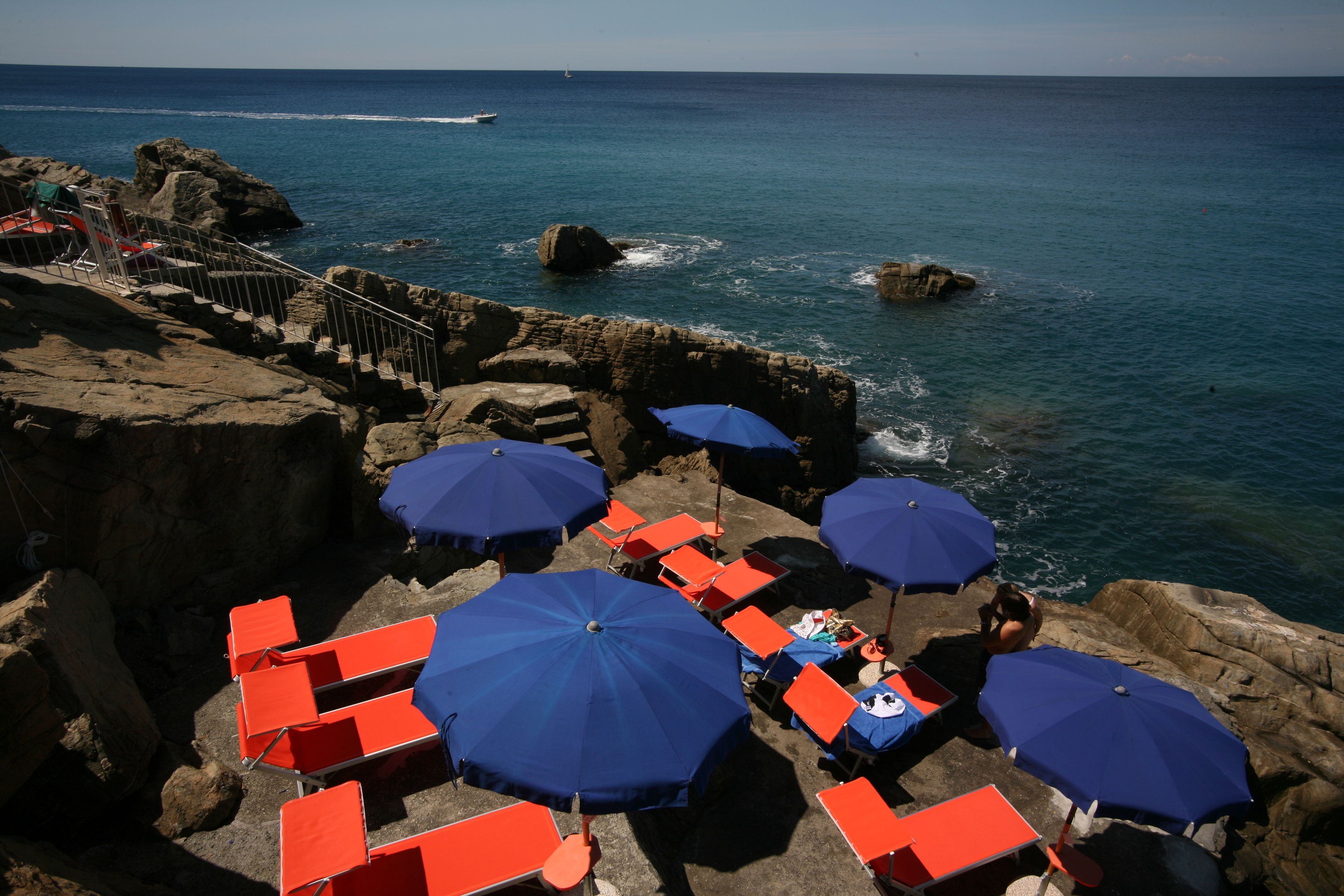 A coastal area with rocky terrain, a staircase leading down to the beach, and several blue umbrellas providing shade.