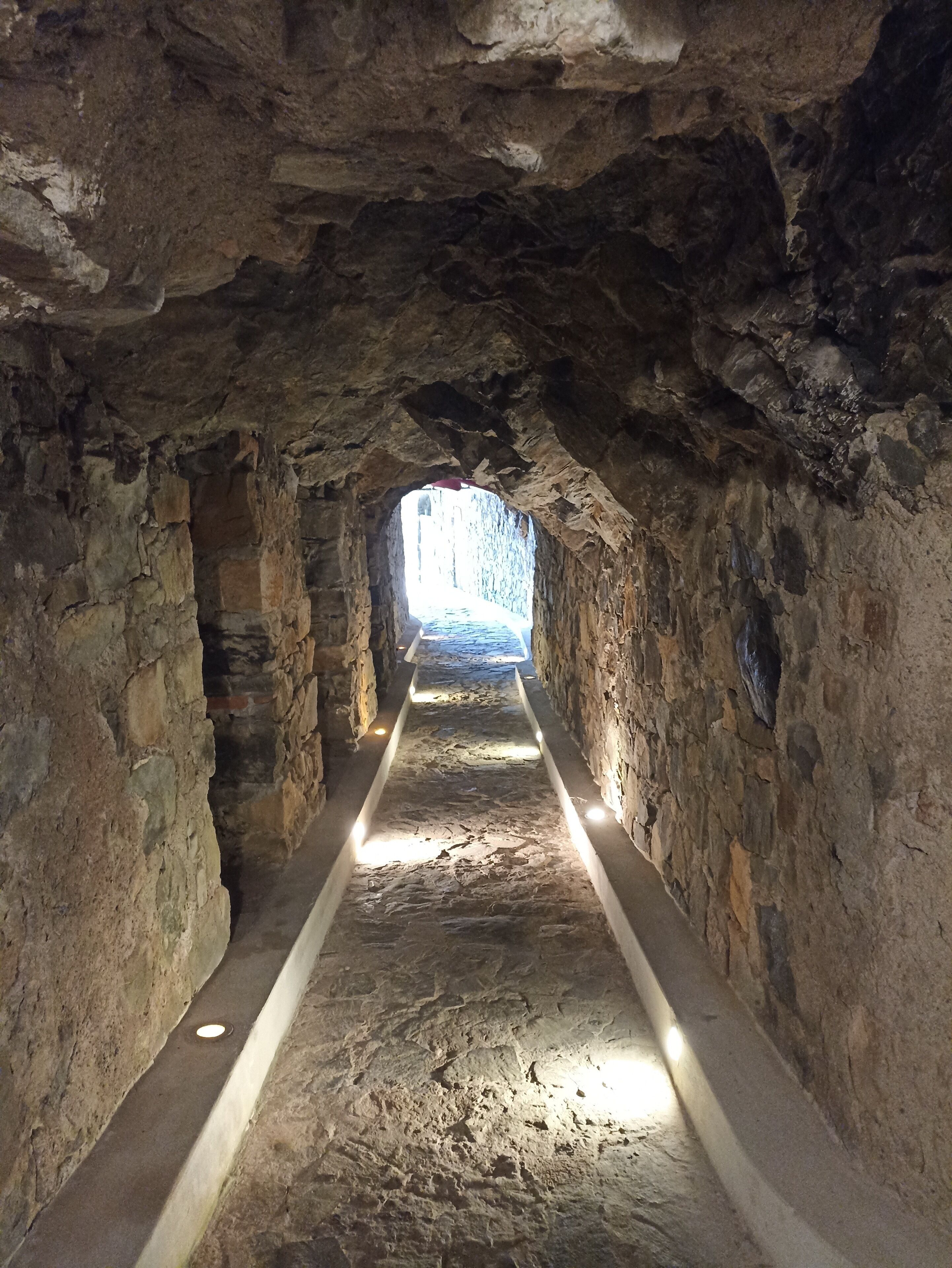 Un estrecho pasaje de piedra con un sendero iluminado en el centro que conduce a una zona más luminosa al final.