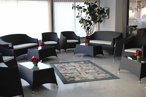 Eine Hotteillobby mit einer zentralen Sitzecke, ausgestattet mit schwarzen Sofas und Sesseln, einem gemusterten Teppich und einer großen Küstpflanze.