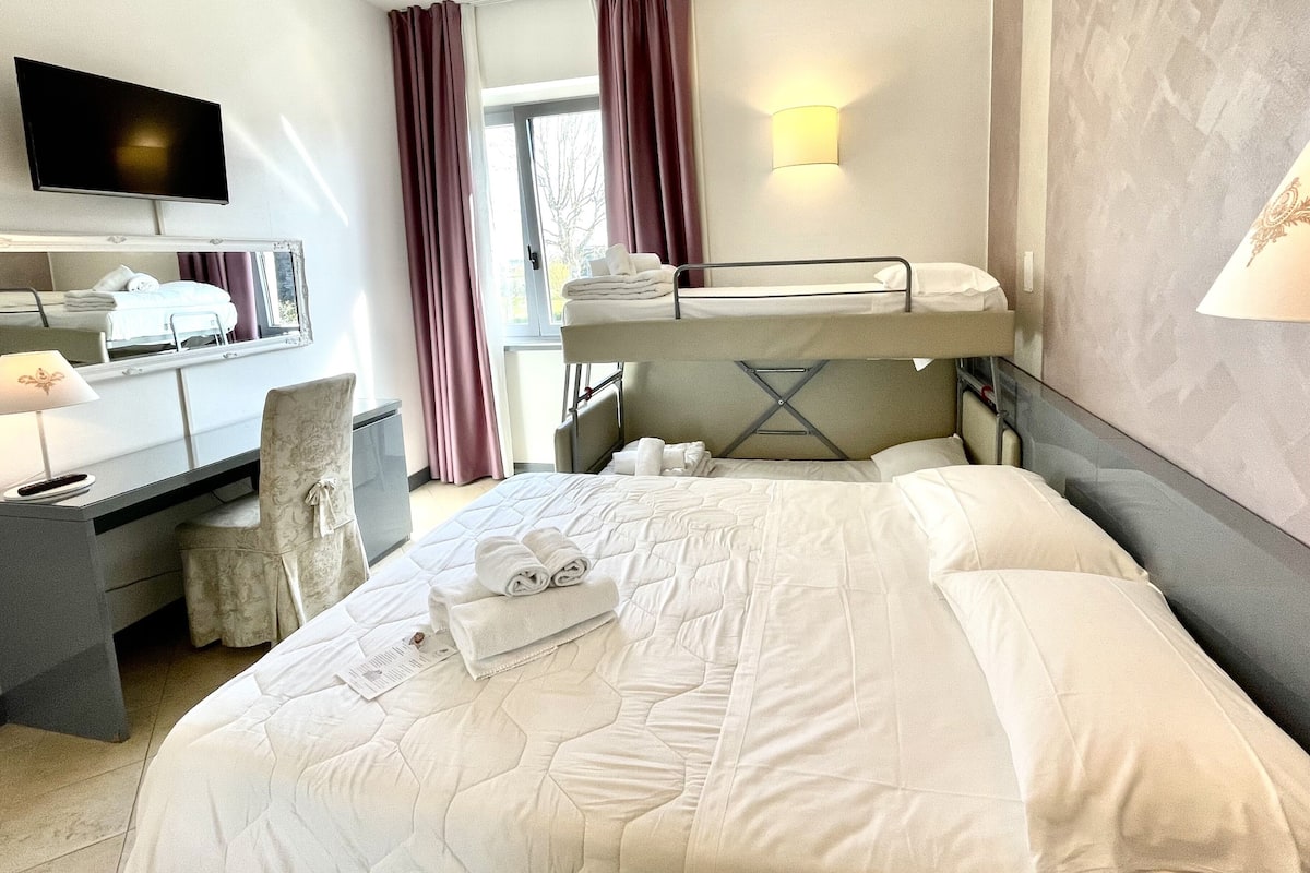 A hotel room with a bunk bed, a desk with a chair, a flat-screen TV, a bedside lamp, and a large window with curtains.