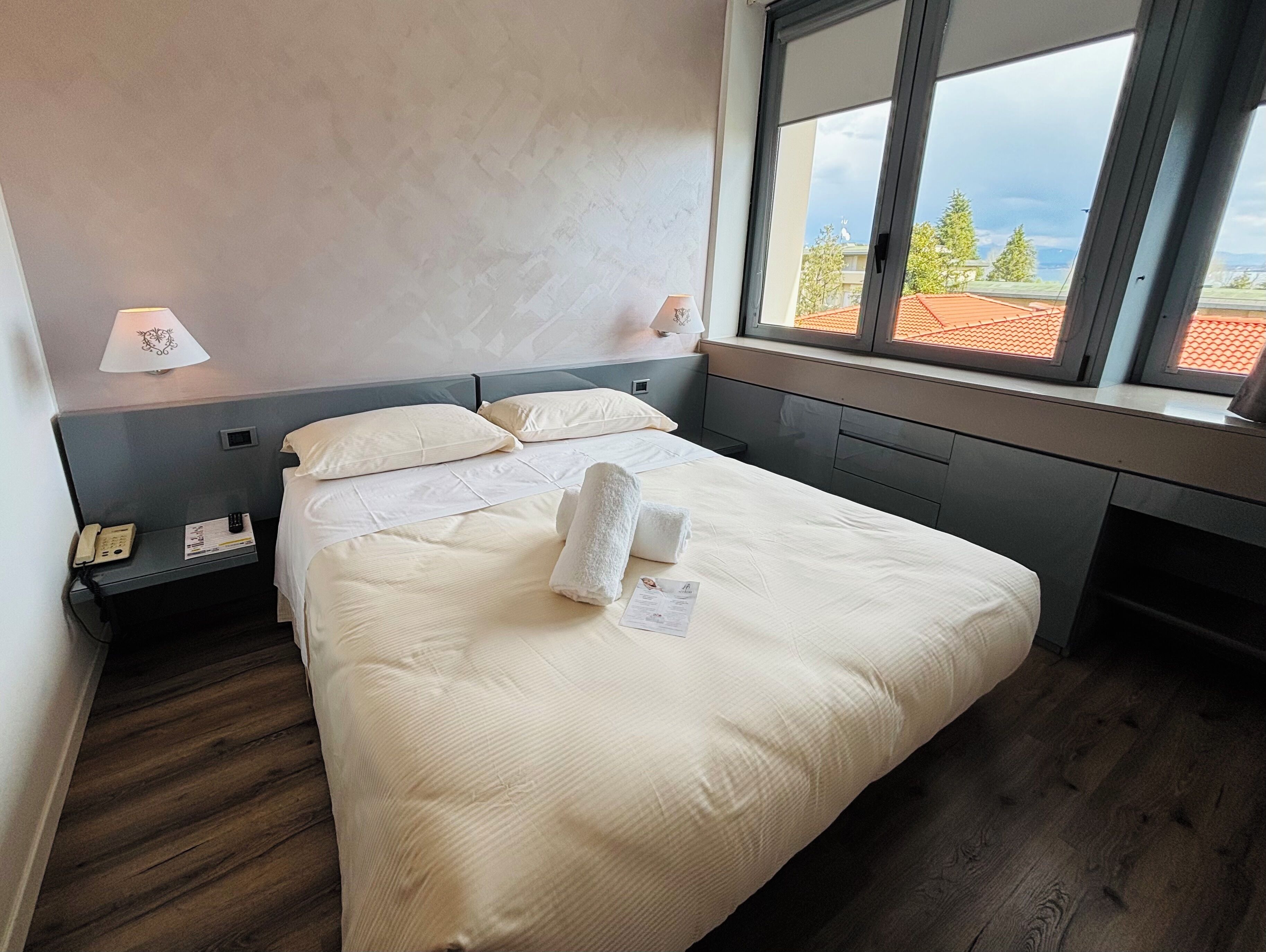 Une chambre d’hôtel avec un lit, une lampe de chevet, un téléphone et une fenêtre donnant sur des immeubles et des arbres.