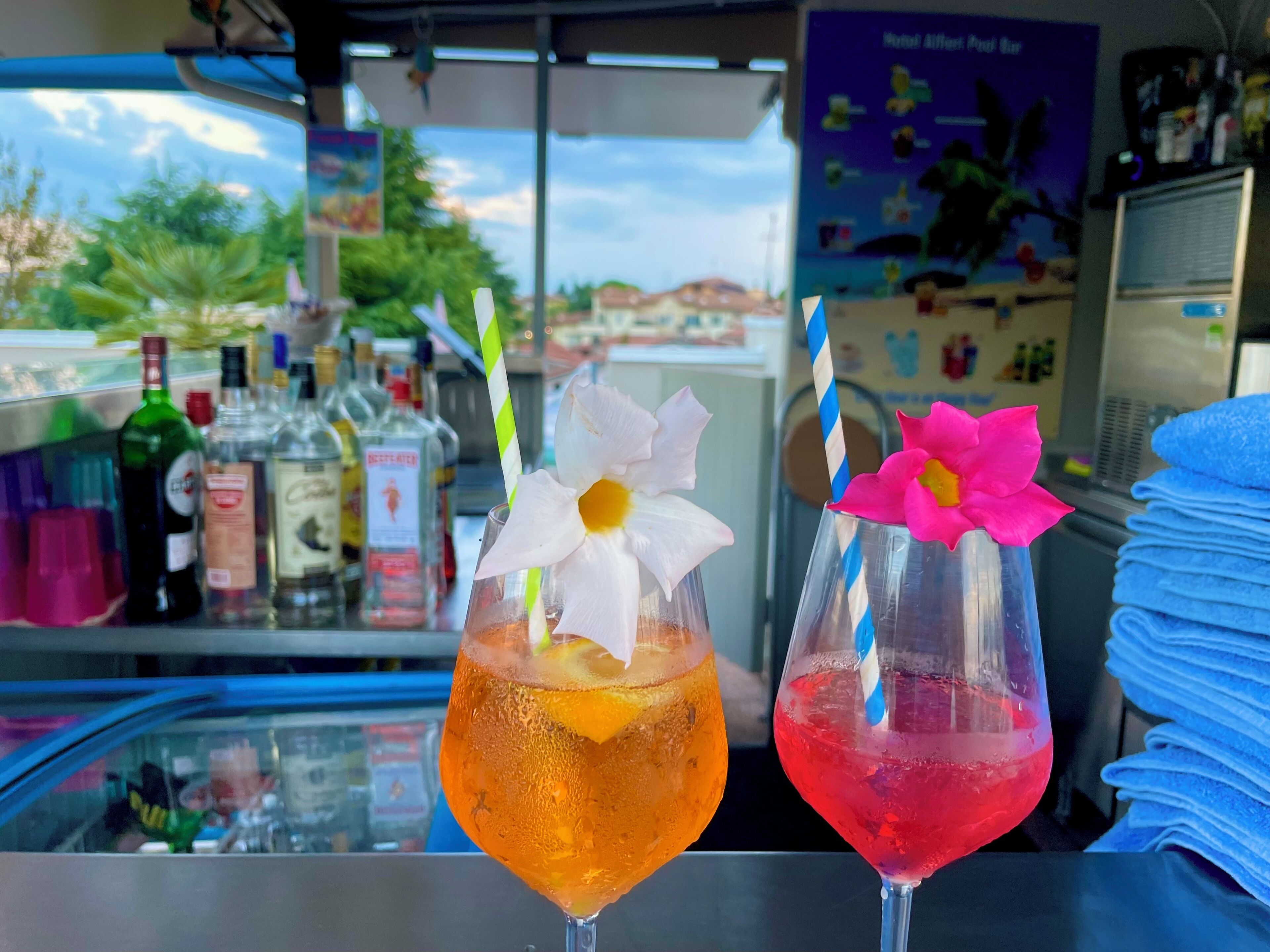 Dos cócteles con adornos florales sobre una barra, rodeados de diversas botellas y un tablero de menú.