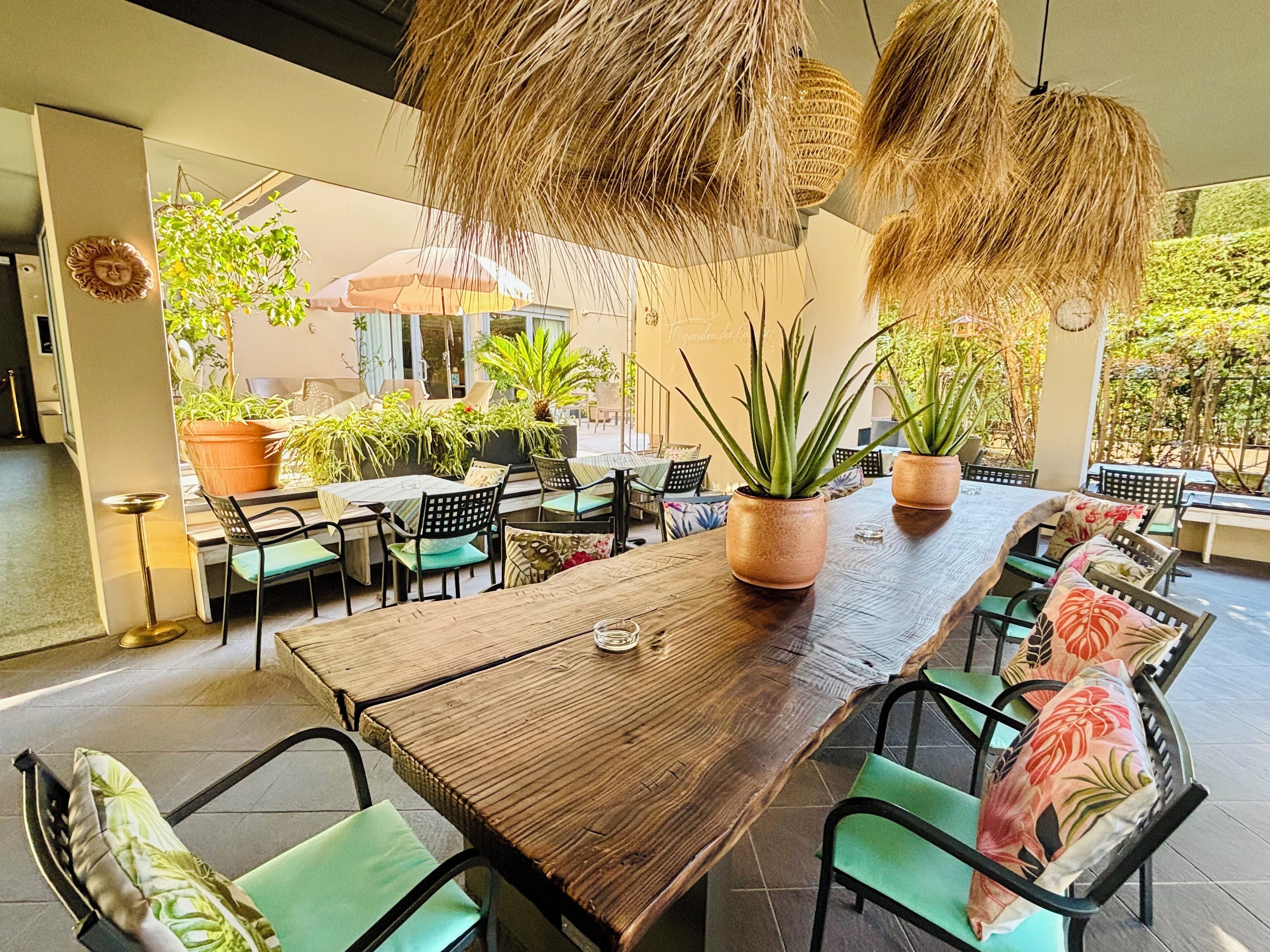 An outdoor dining area with wooden tables and chairs, potted plants, and hanging thatch lamps.