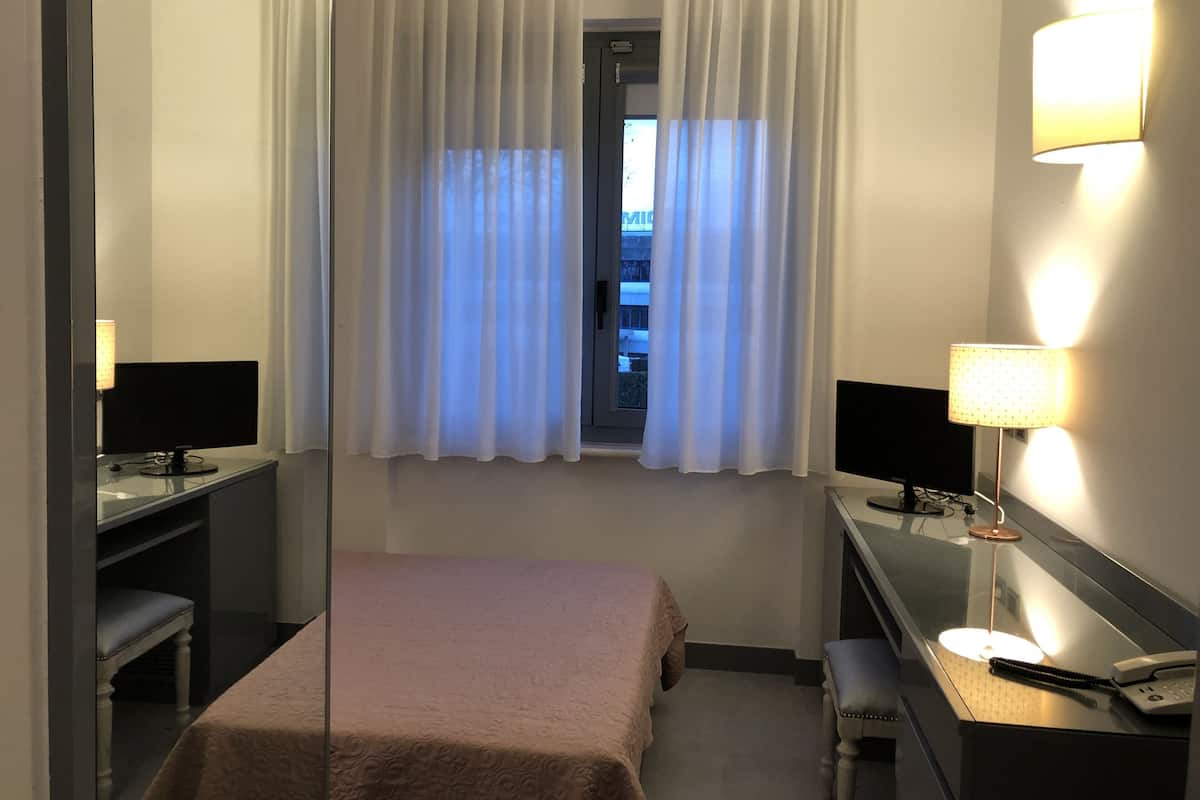 A hotel room with a bed, a desk with a computer, a chair, a lamp, and a window with curtains.