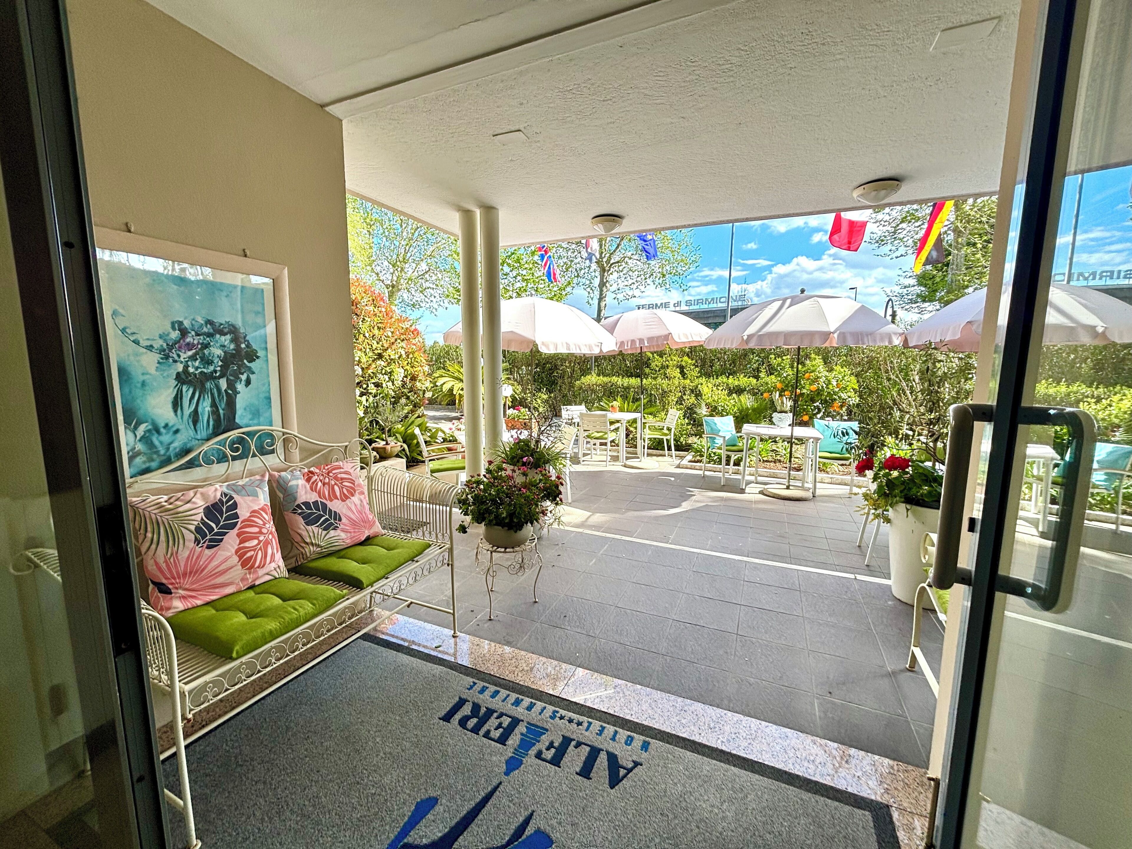 A hotel entrance with a reception desk, a seating area with a sofa and chairs, and a view of the outdoor garden and umbrellas.