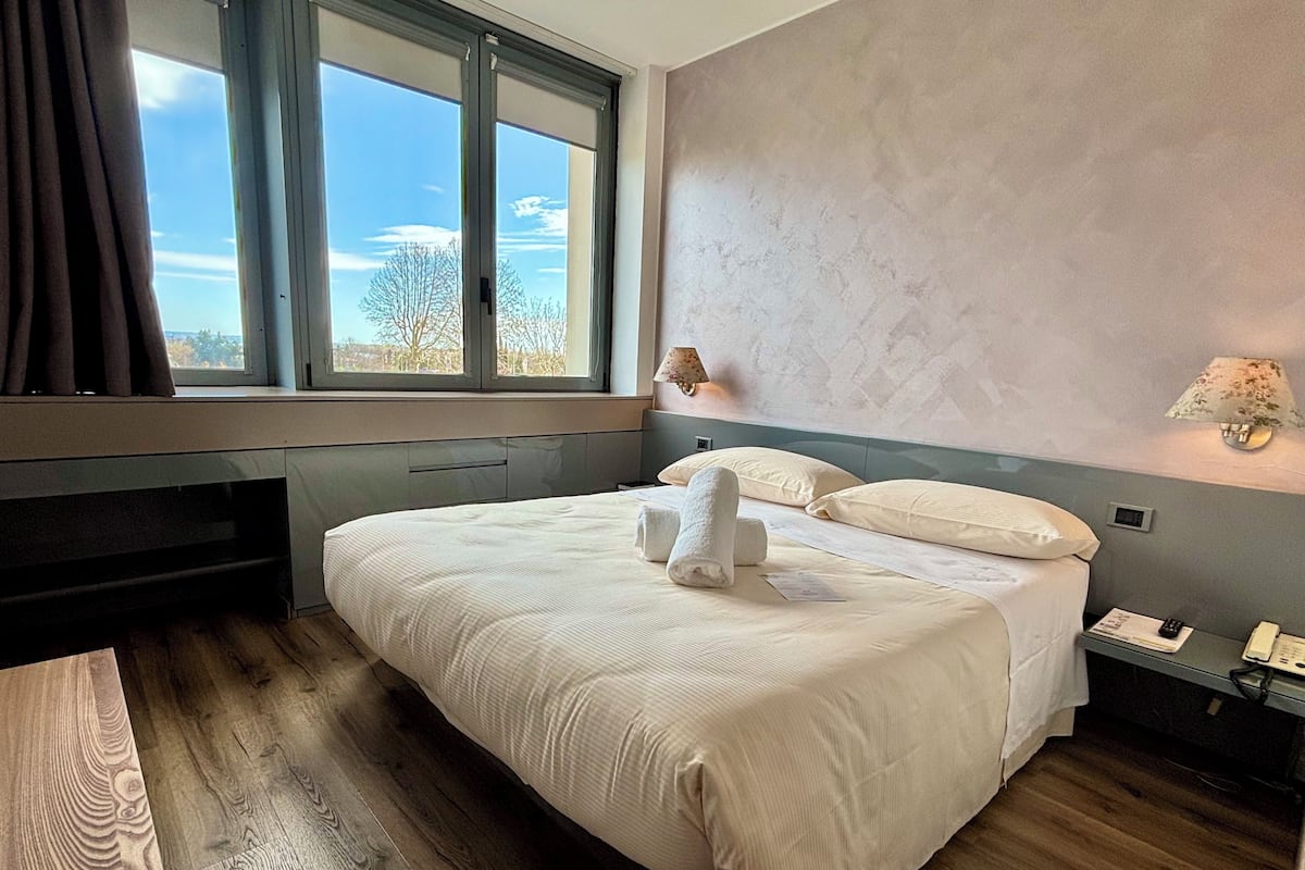 A neatly made bed with white linens and two towels, a bedside table with a lamp, and a view of the outdoors through a large window.