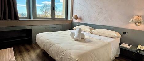 A neatly made bed with white linens and two towels, a bedside table with a lamp, and a view of the outdoors through a large window.