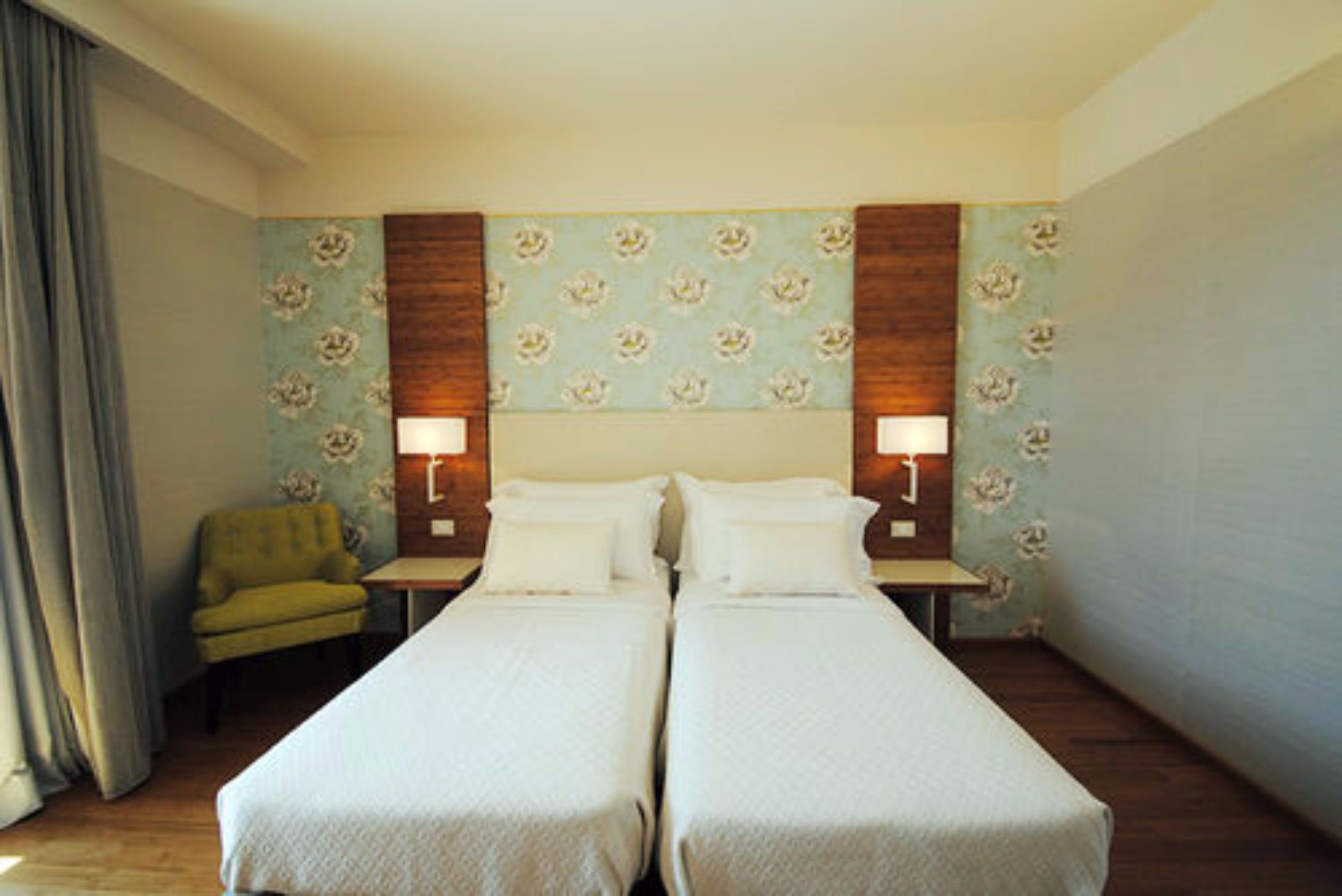 Una habitación de hotel con dos camas individuales, un sillón pequeño y dos lámparas de noche.
