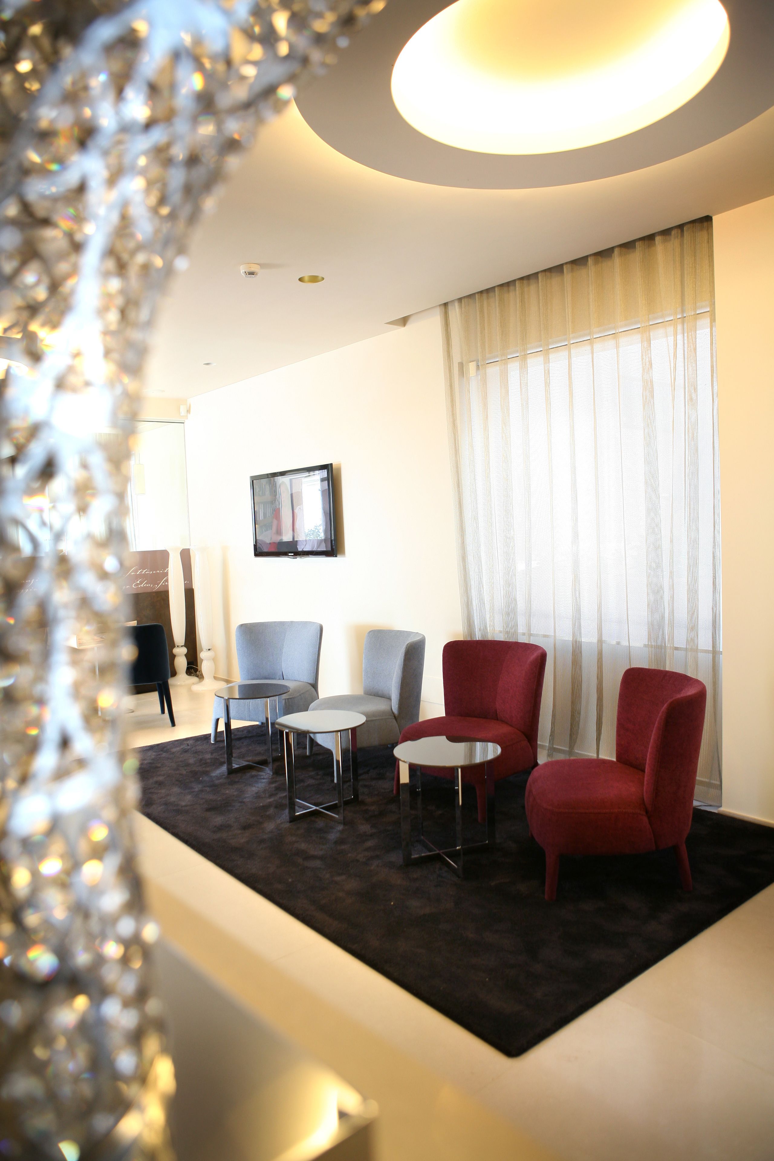 A modern hotel room with a dark carpet, two red armchairs, a grey armchair, a small round table, and a television on the wall.