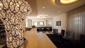 A modern hotel lobby with a central decorative column, seating area, and a reception desk. - Hotel Eden (Sirmione)