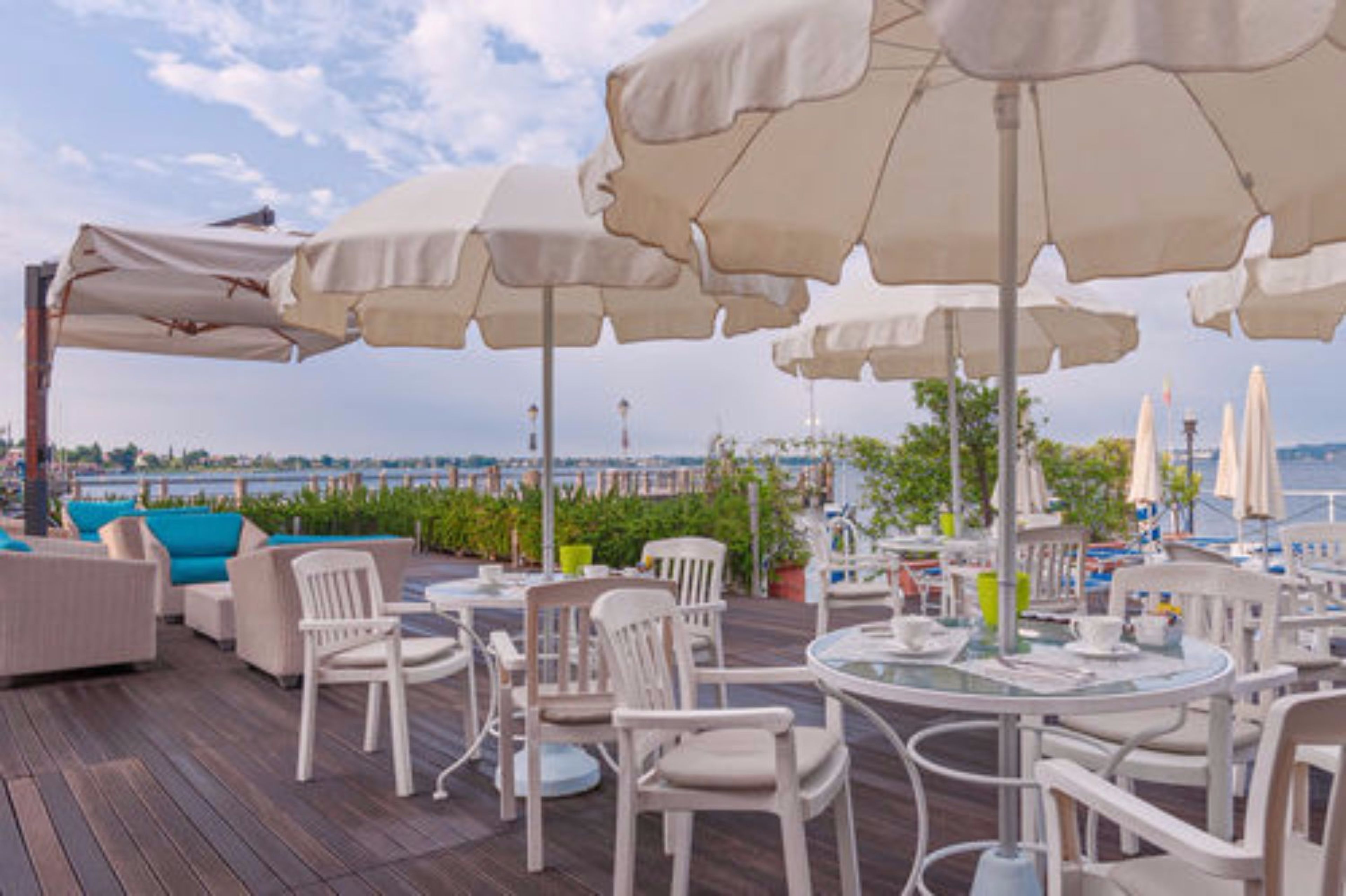 An outdoor seating area with white chairs and tables, beige umbrellas, and a view of the water.
