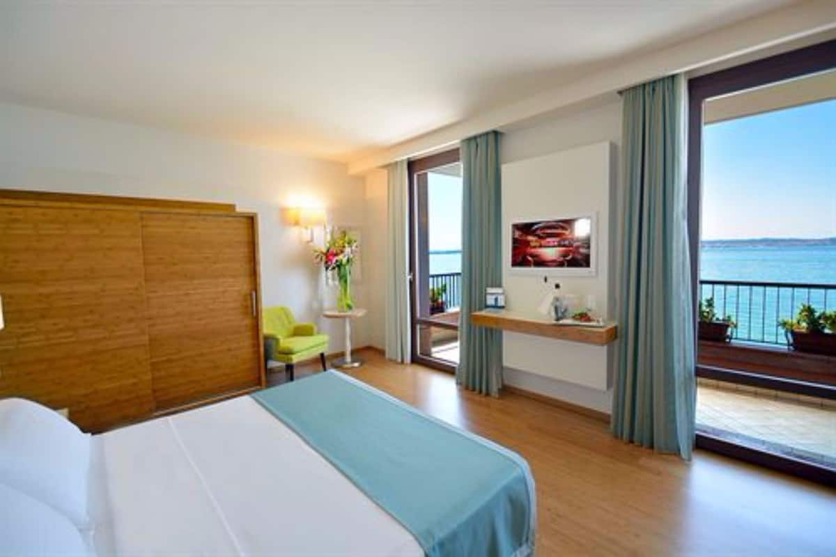 Een hotelkamer met een bed, een stoel, een klein tafeltje met een bloevas, een televisie op een standaard en een balkon met uitzicht op de zee.
