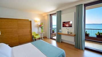 Una camera d'albergo con un letto, una sedia, un tavolino con un vaso di fiori, una TV su un mobile e un balcone vista mare.