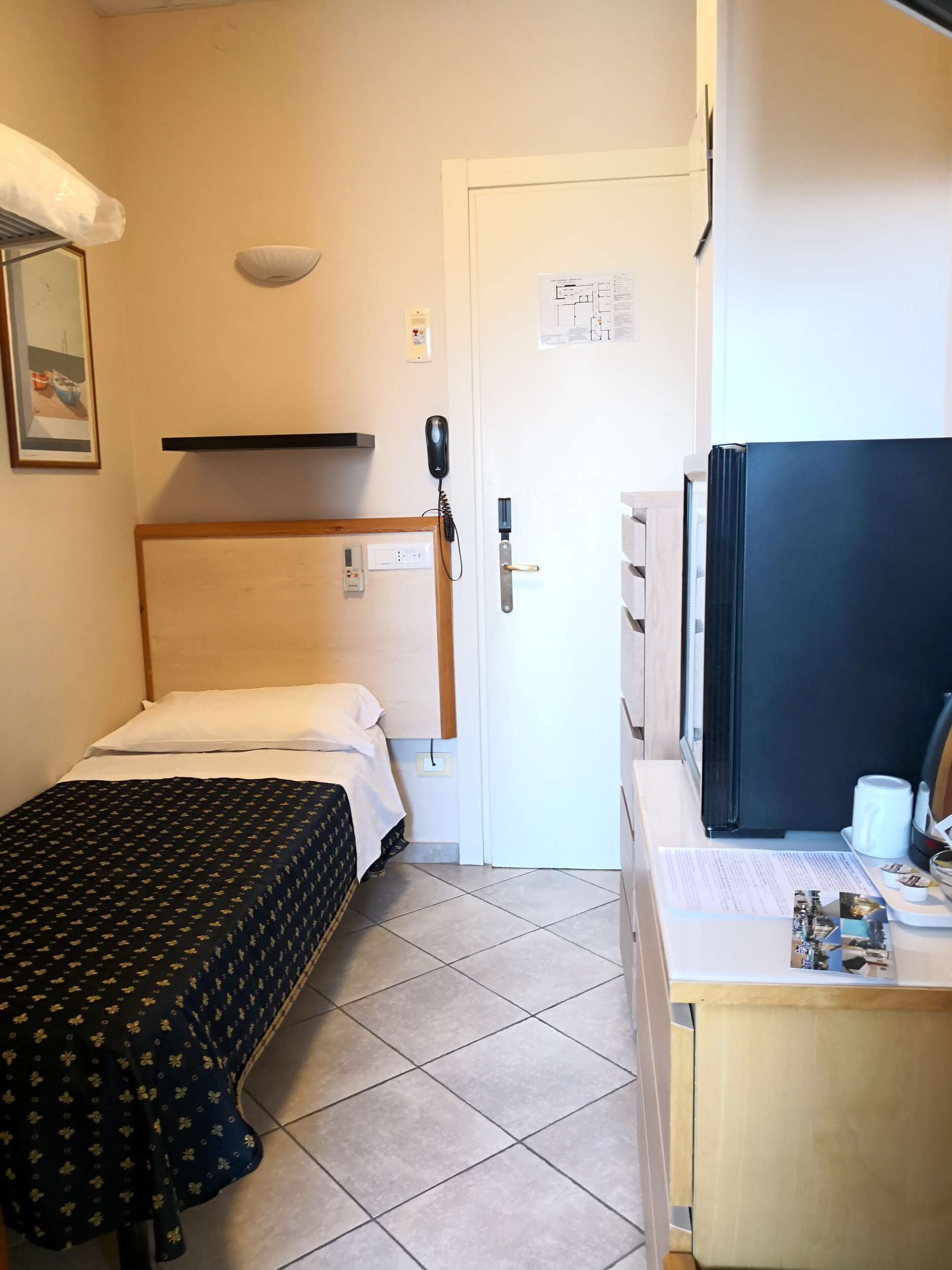 Una stanza piccola e pulita con un letto, una scrivania con un computer e un frigorifero.