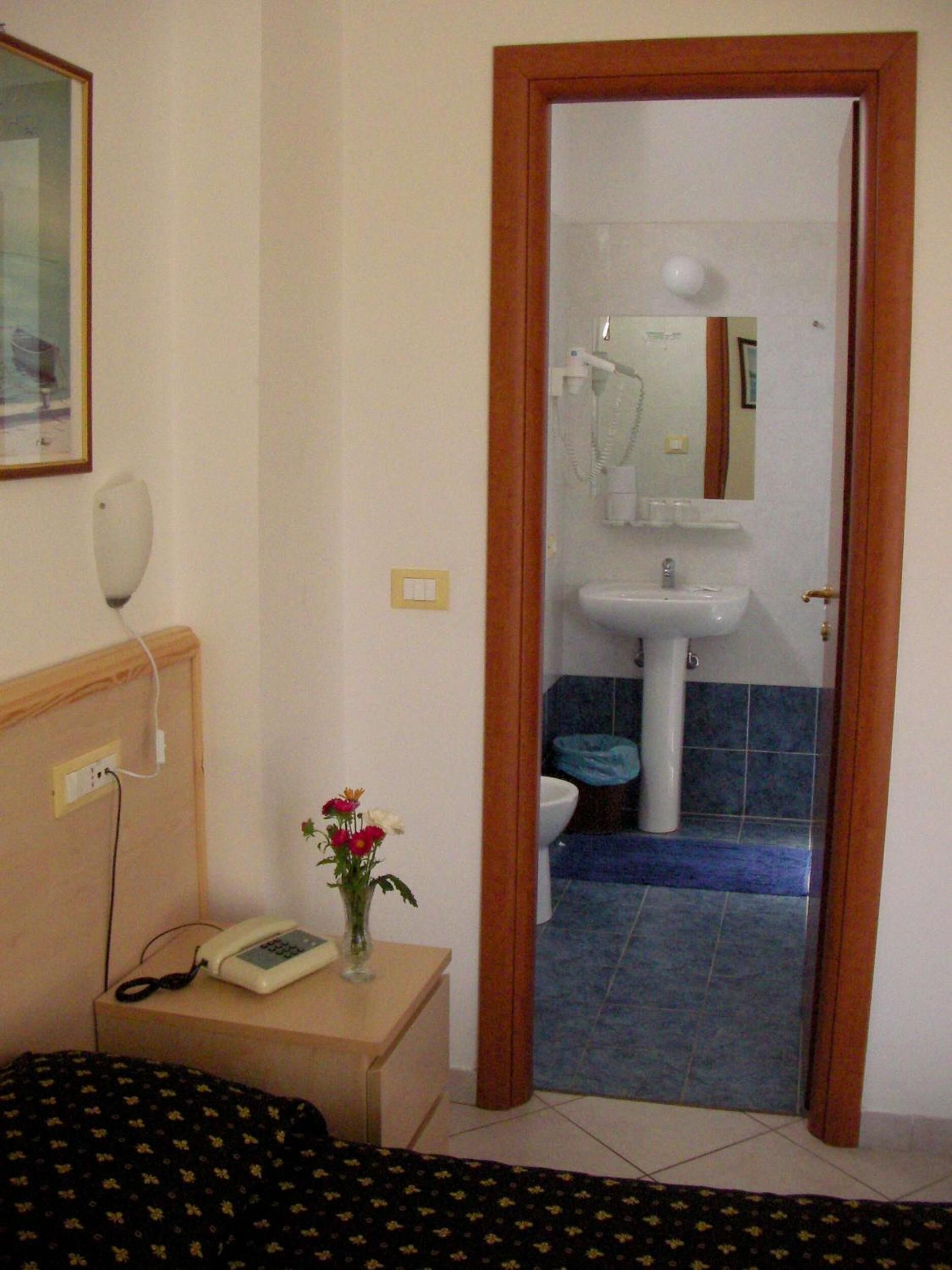 Et hotelværelse med en seng, et natbord med telefon og vase, et lille bord med blomster og et badeværelse, der kan ses gennem en åben dør.